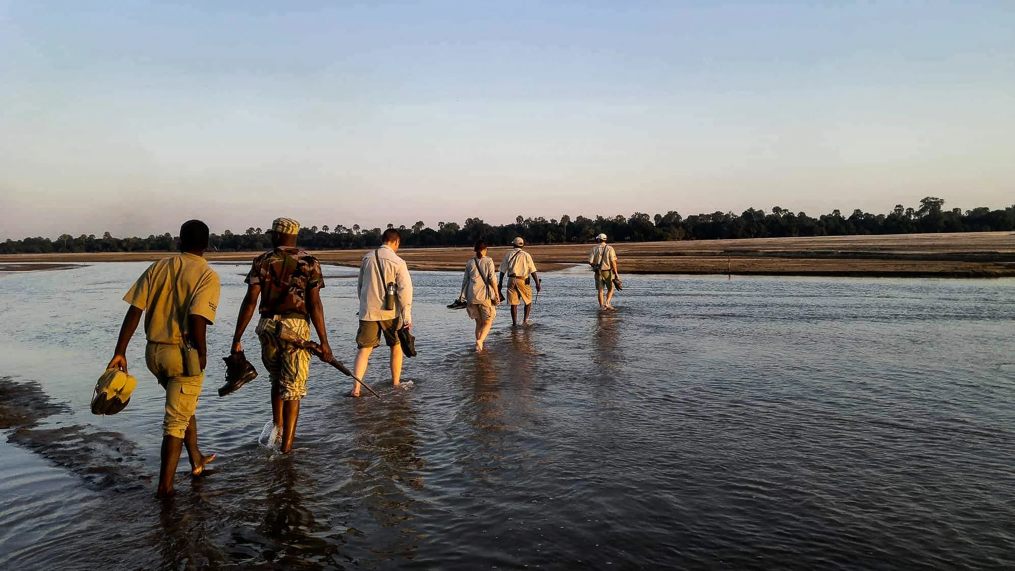A group of seven people walking through shallow water on a riverbank at sunset, with a treeline in the distance.