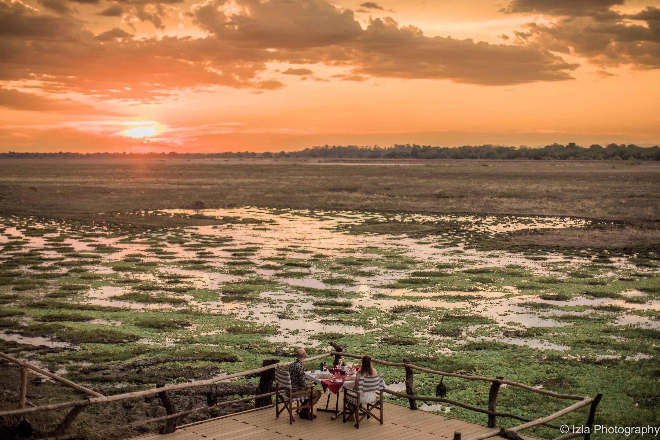 A couple dining outdoors at a wooden table on a deck overlooking a swamp at sunset, with water and vegetation under a partly cloudy sky.