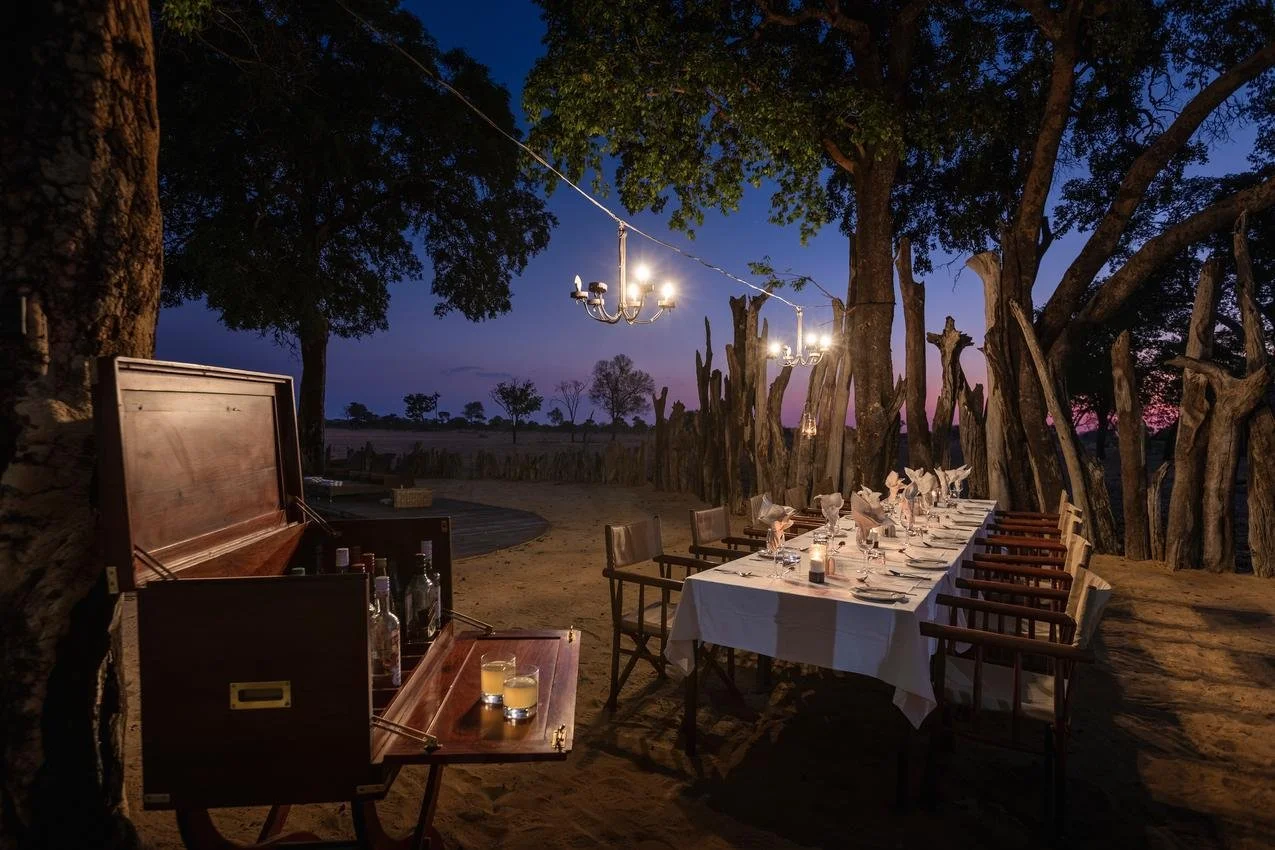 An outdoor dining setup at dusk with a long table set with glasses and napkins, surrounded by wooden chairs, under trees with a string of chandeliers hanging above, on a sandy ground in a rustic setting.