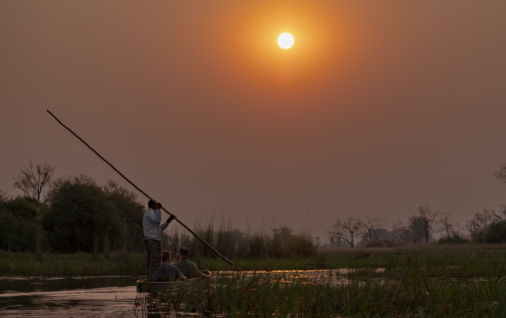 Three people in a boat on a river at sunset, with one person standing and holding a long pole, and two others sitting, surrounded by trees and tall grasses.