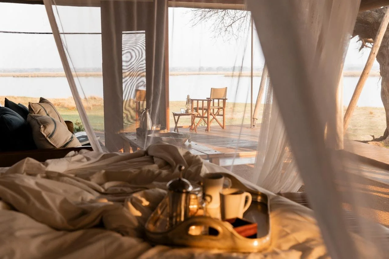 A cozy bed with a tray holding mugs, a teapot, and a wooden box, looking out onto a porch with chairs and a view of a lake or river at sunset.