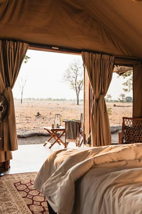 Inside a tent with an open flap looking out onto a flat, dry landscape with sparse trees. There is a bed with white bedding in the foreground and a small wooden table outside with a lantern, a cup, and a blanket draped over a chair.
