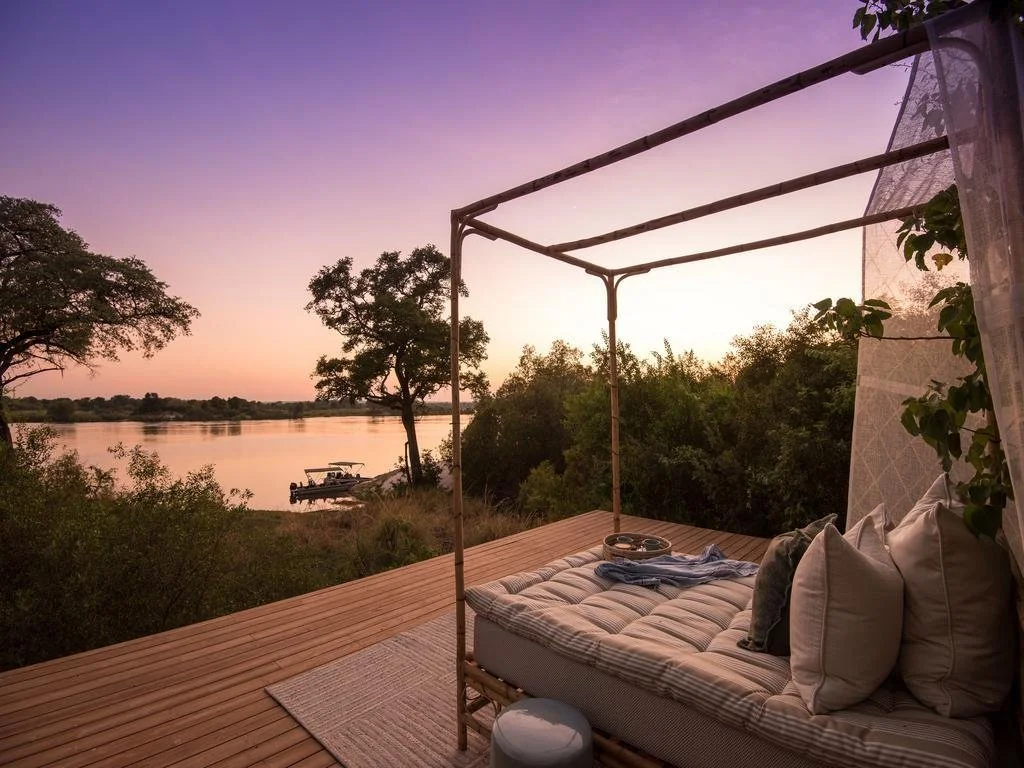 Sunset view over a river with a boat docked on the shore, seen from a cozy outdoor lounging area with a bed, pillows, and a canopy frame on a wooden deck.