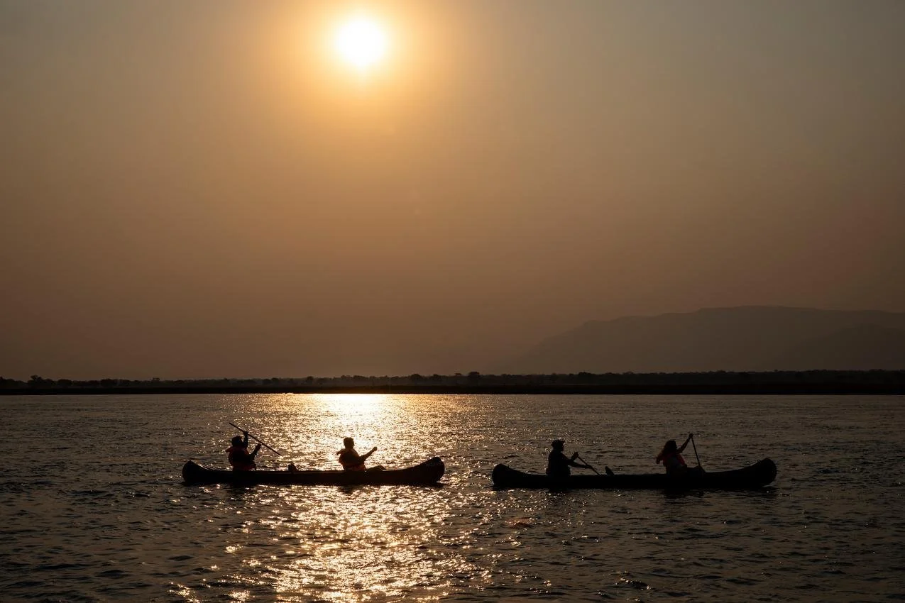 Four people kayaking on a calm body of water during sunset with silhouetted mountains in the background.