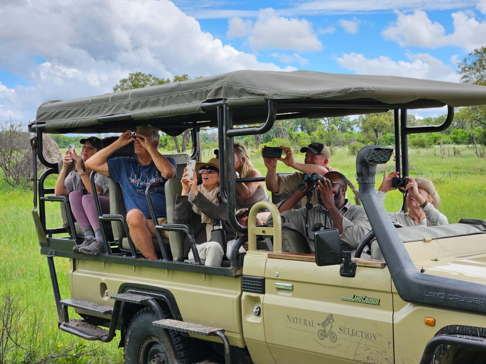 Tourists sitting on a safari vehicle in a grassy field taking photos of wildlife.