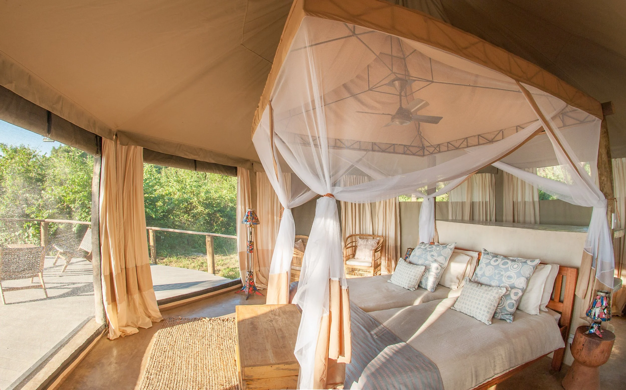 Interior of a bedroom with a four-poster bed featuring a white canopy and patterned pillows, large windows with beige curtains, and outdoor seating with chairs and a table visible through the windows, surrounded by greenery.