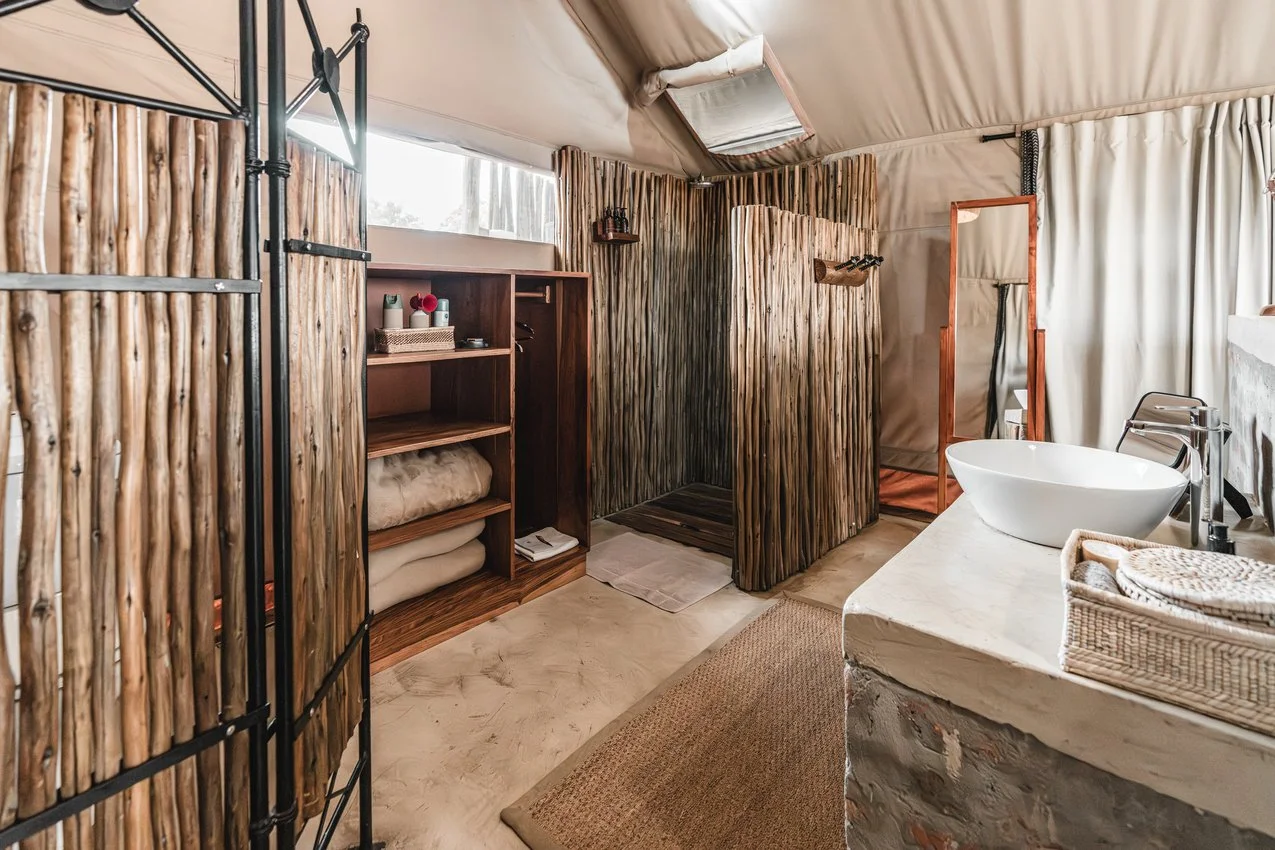 Rustic bathroom with wood-paneled dividers, a standing mirror, sink with a woven basket, and a shower area enclosed by wood-like panels, inside a tent-like structure with fabric ceiling and walls.