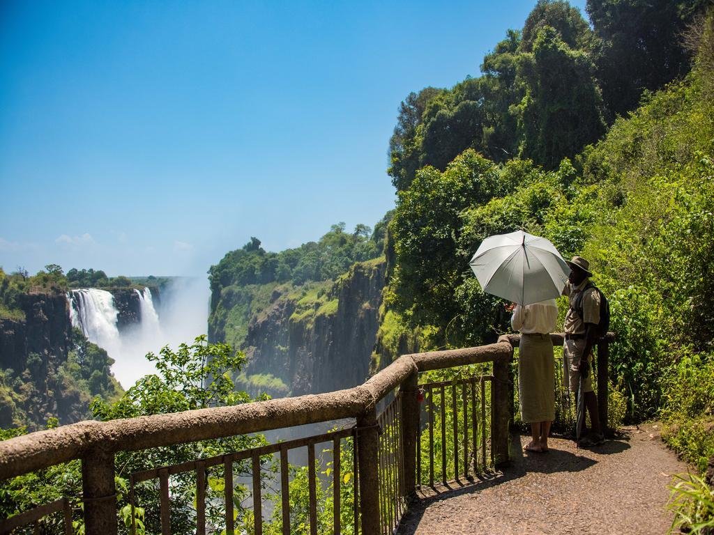 Two people, one holding a white umbrella, standing on a viewing platform overlooking a waterfall surrounded by lush green trees under a clear blue sky.