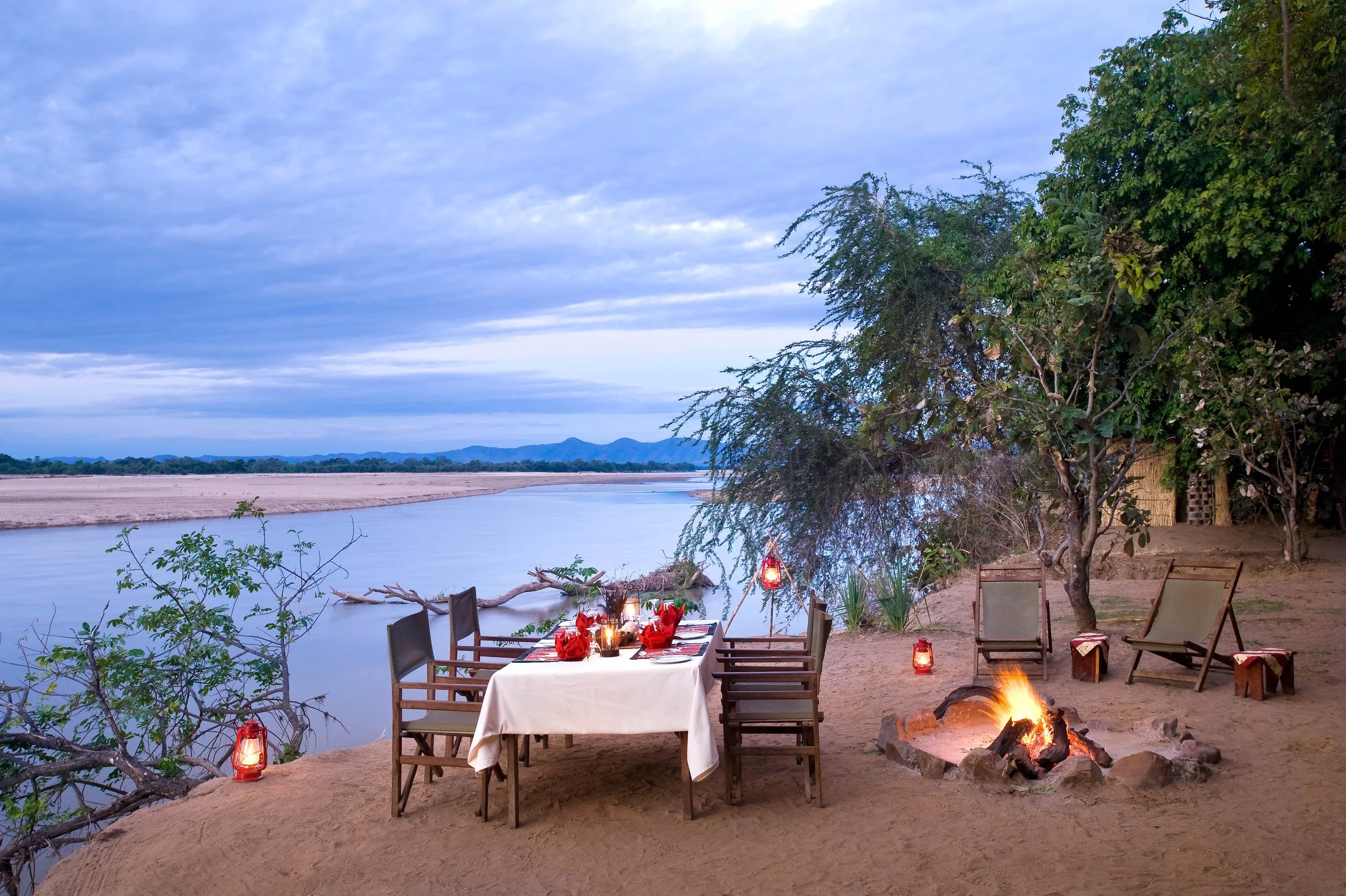 Dinner table set up outdoors by a river with chairs, lanterns, and a campfire, surrounded by trees under a cloudy sky.