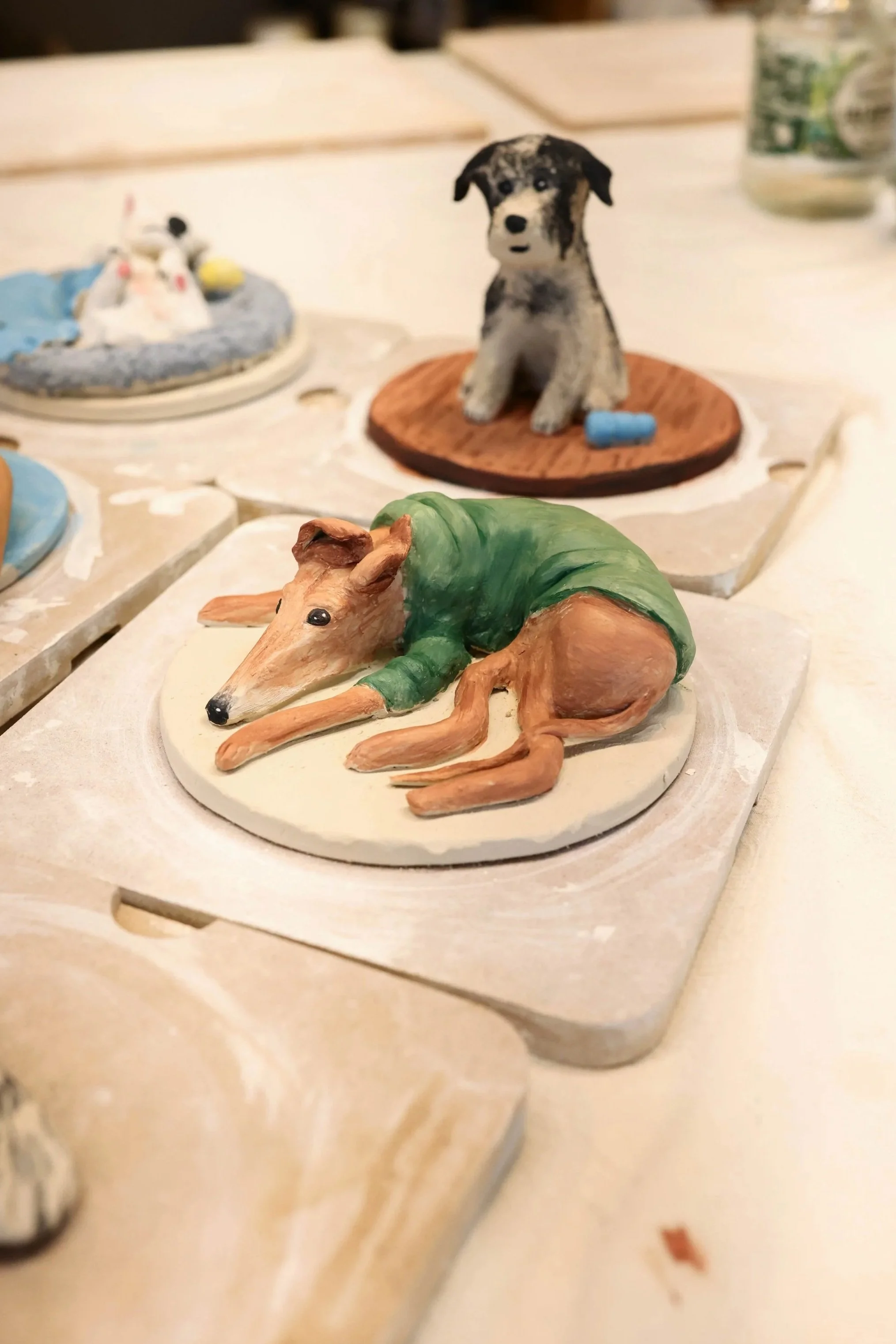 A woman working on a clay sculpture in an art studio, surrounded by small ceramic animal figurines and art supplies such as brushes, a jar, and a container with pink clay or dough.