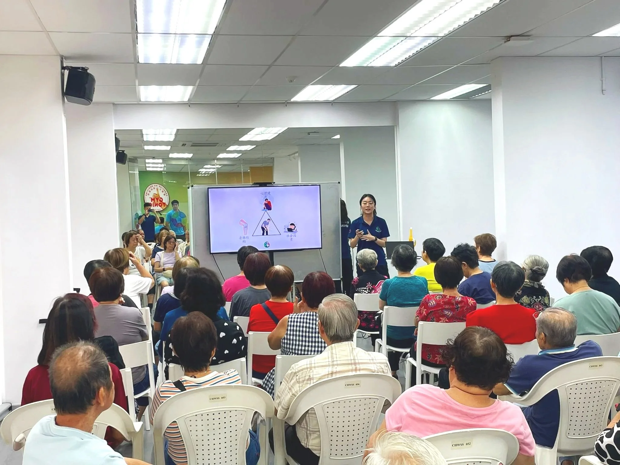 A woman presenter standing next to a large display screen showing a diagram, addressing an audience of mostly elderly people seated in chairs at an indoor event.
