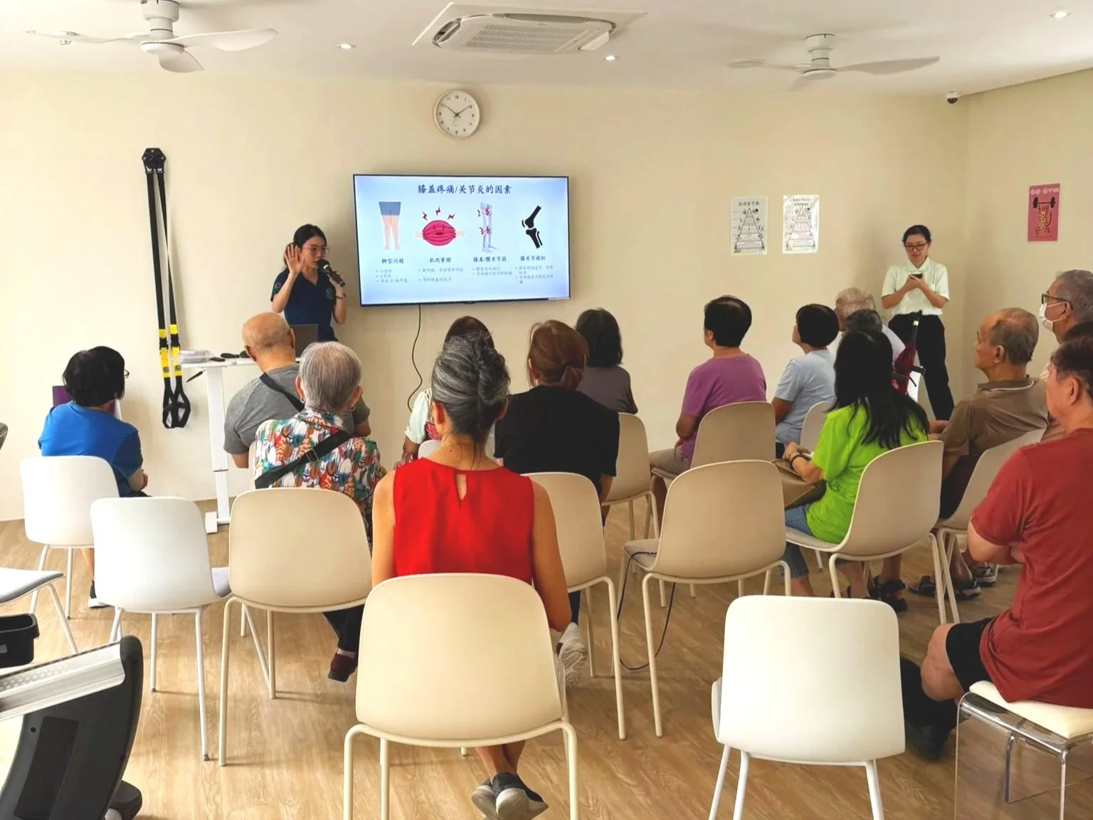 A group of people attending a health presentation in a well-lit room with a large screen displaying information about joint health, with a woman speaking at the front and an instructor standing nearby.