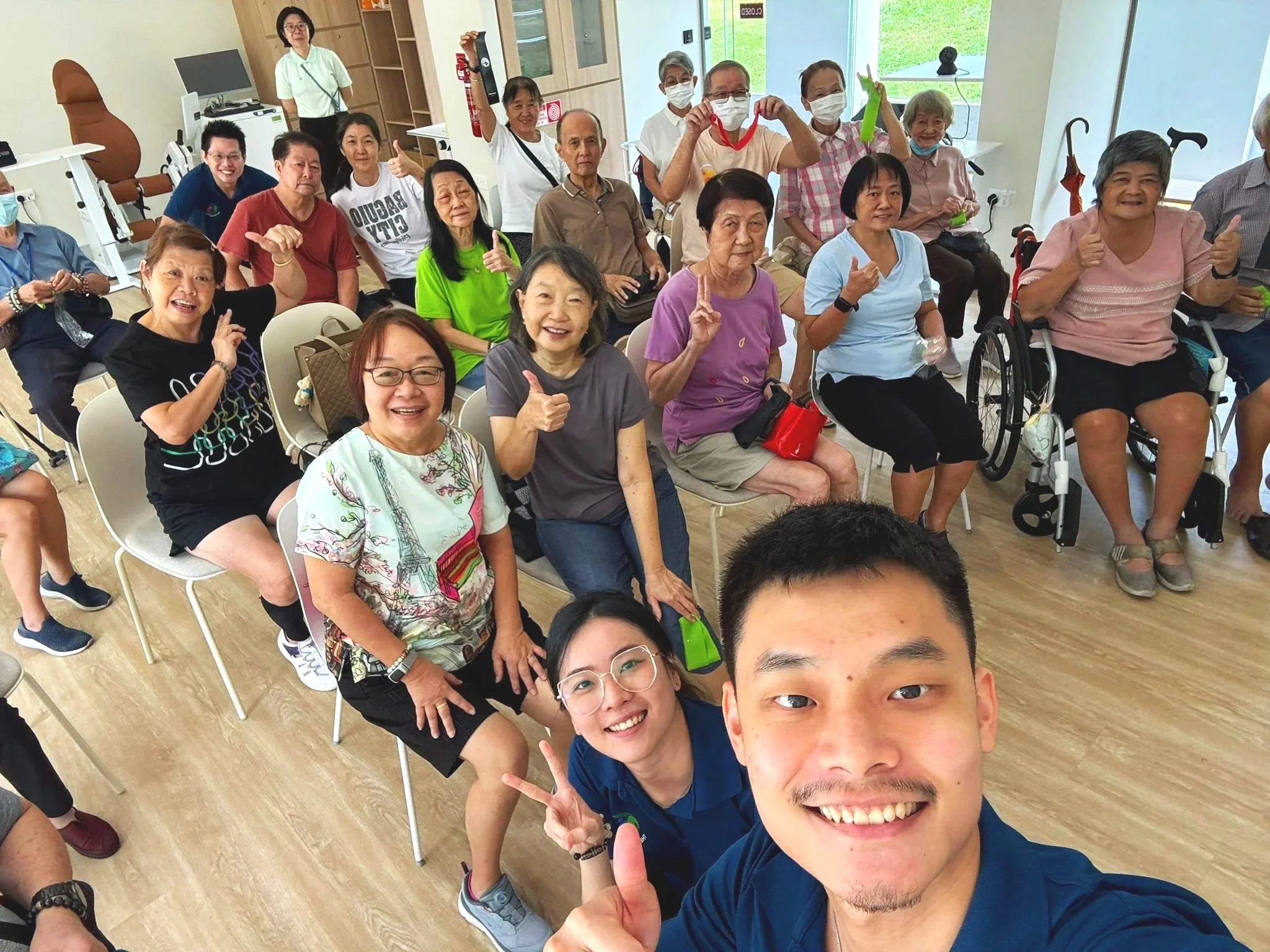 Group of elderly and middle-aged people, some in wheelchairs, seated and smiling in a bright room during a social gathering or activity. Two young men are taking a selfie in front of the group, smiling and making peace signs.