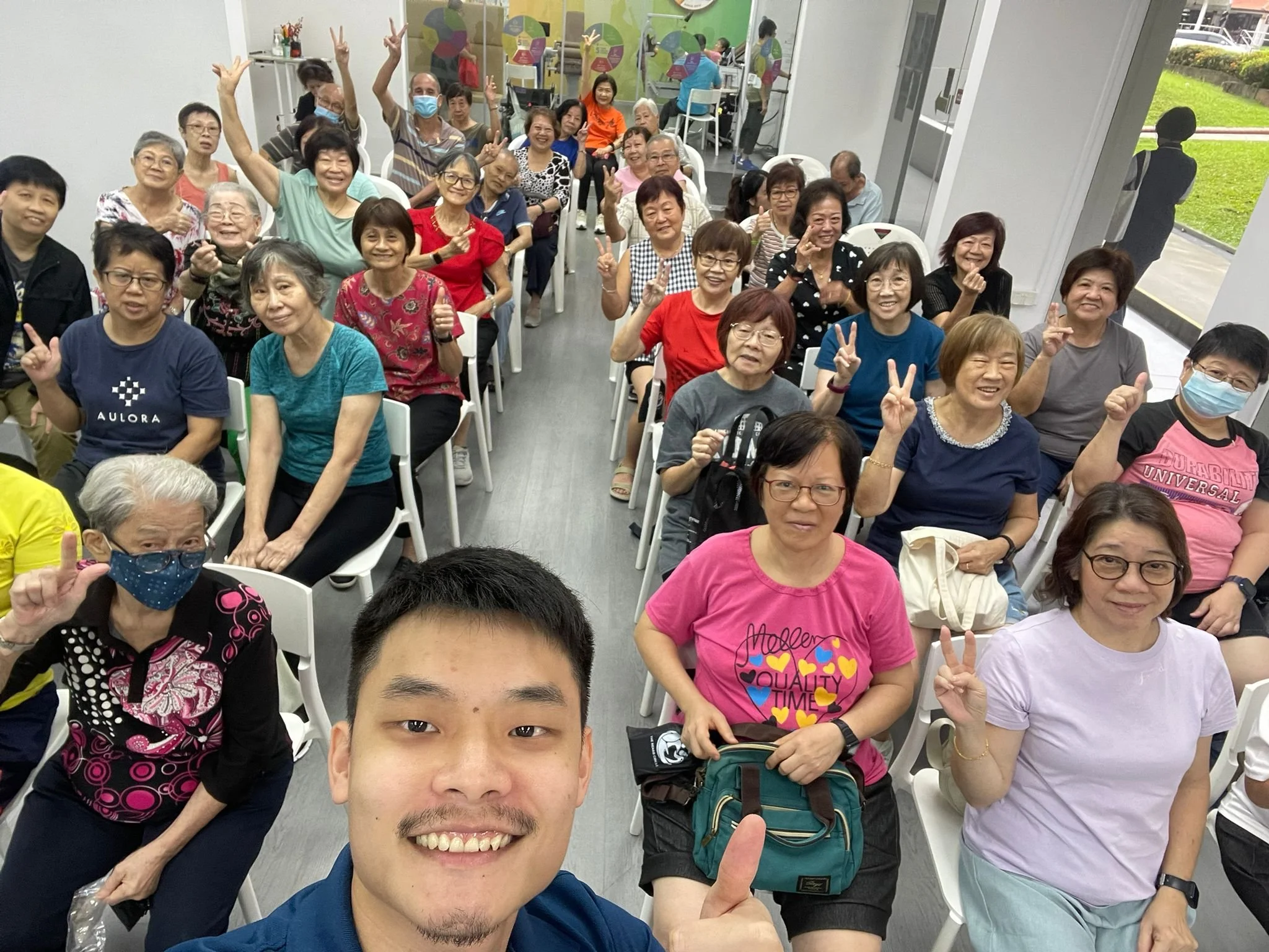 A group of diverse elderly people attending a community gathering indoors, smiling and making peace signs for a selfie taken by a young man in the foreground.