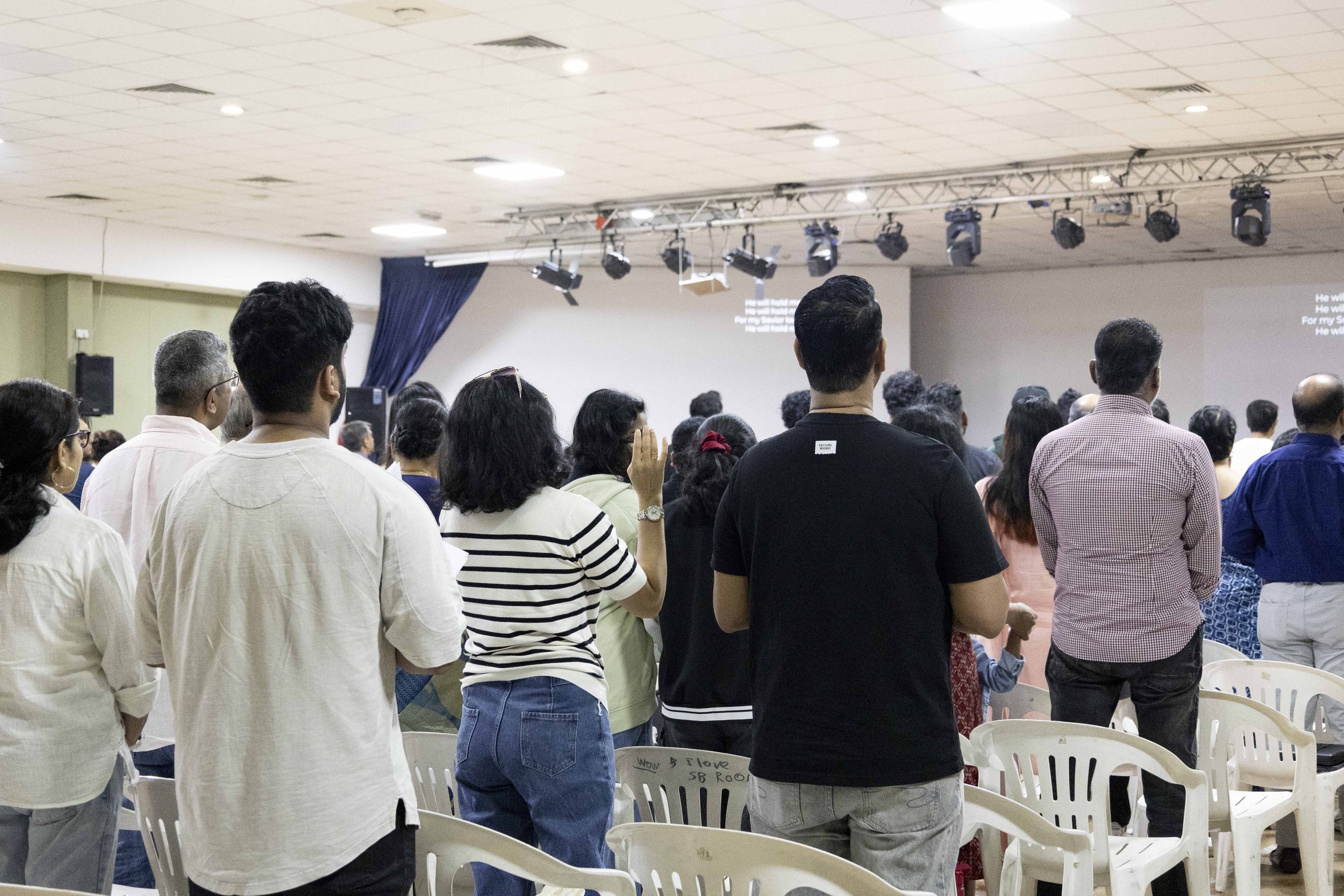 People standing and facing forward in a large indoor auditorium or hall, likely attending a religious or community event. The space has a stage with lighting and a screen at the front.