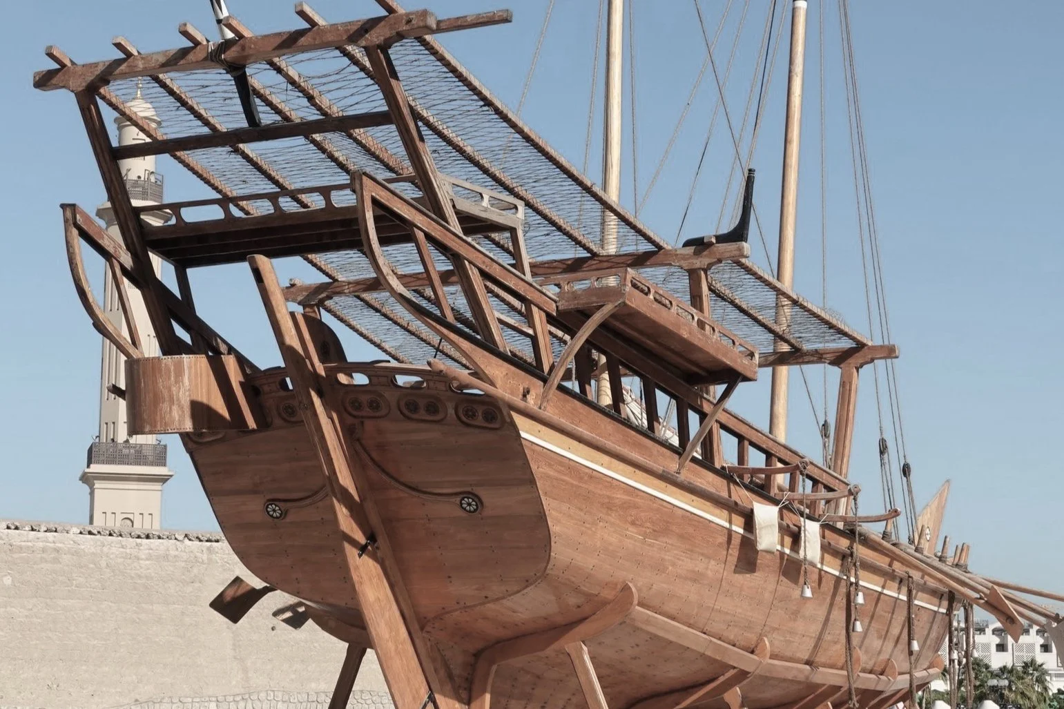 A large wooden ship with masts and rigging, under construction or on display, with a lighthouse in the background.
