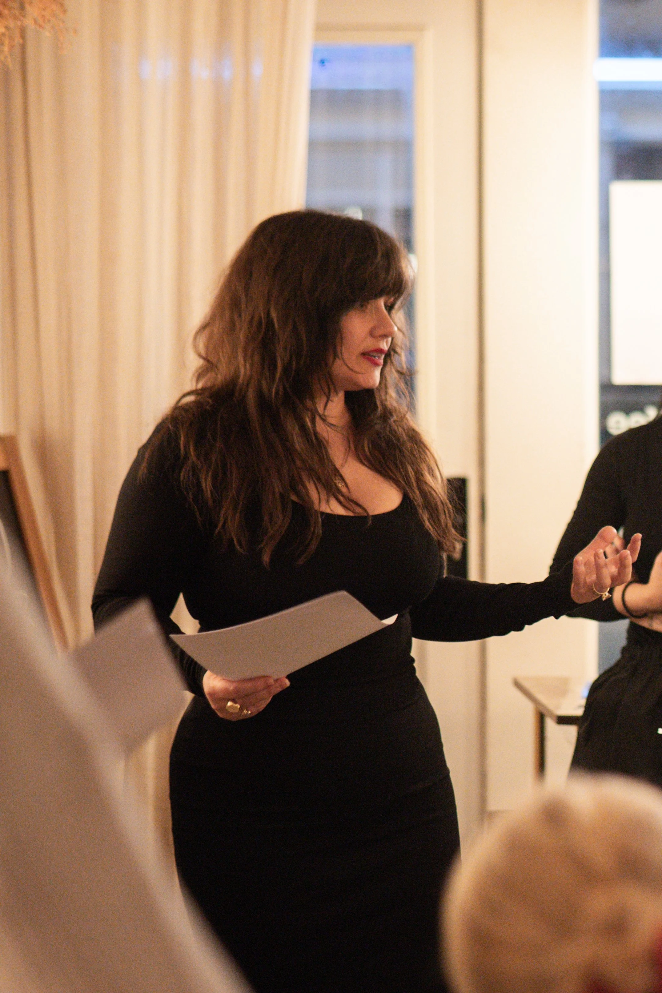 A woman with long brown hair, wearing a black dress, holding a piece of paper, and speaking or presenting in an indoor setting with beige curtains and a window in the background.