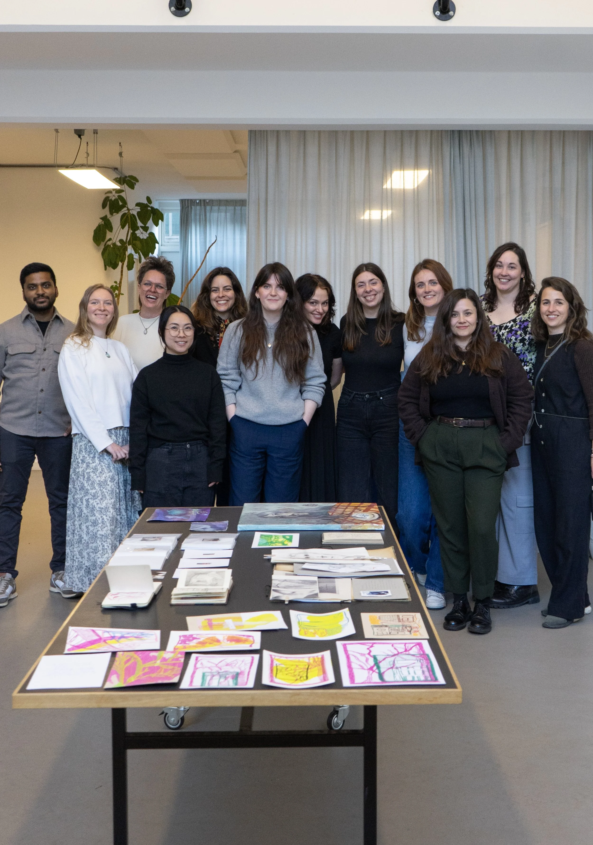 A group of twelve diverse people standing behind an art display table, which holds colorful drawings and sketches, in an indoor setting with white curtains and a large leaf plant in the background.