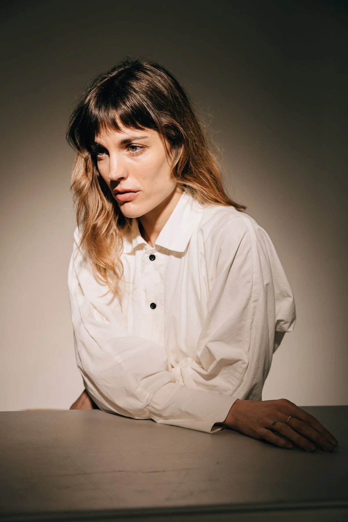 A woman with shoulder-length wavy hair in shades of brown, wearing a white button-up shirt, sitting at a table with her left arm resting on it, photographed against a plain, dark background.