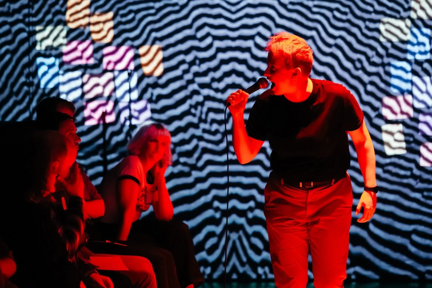 A person with short blonde hair singing into a microphone on stage with a black background and a zebra stripe pattern projected behind them. Two people are seated in the foreground, watching the performance.