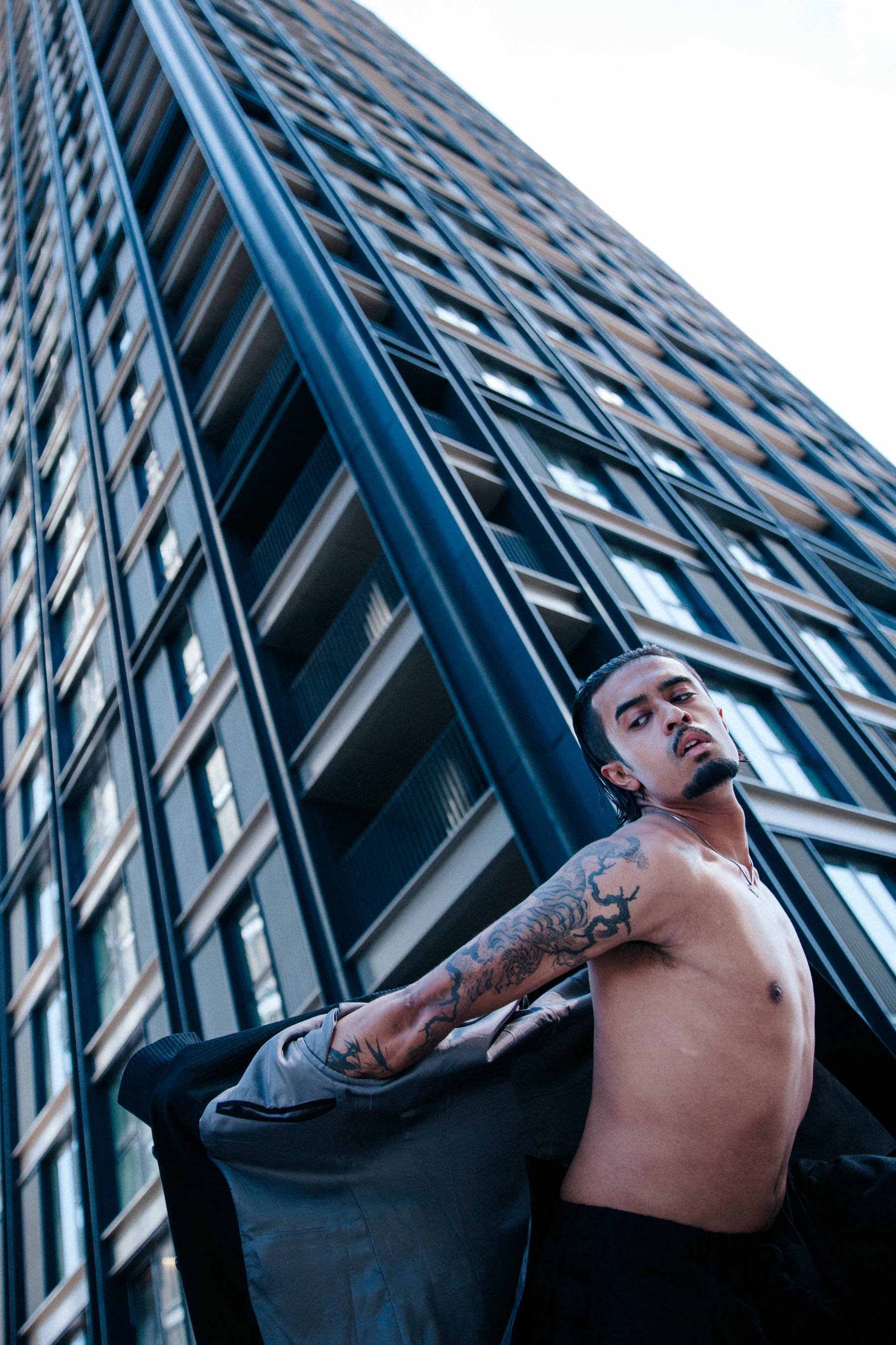 A shirtless man with tattoos on his arm and chest stands against the backdrop of a tall modern building, looking at the camera.