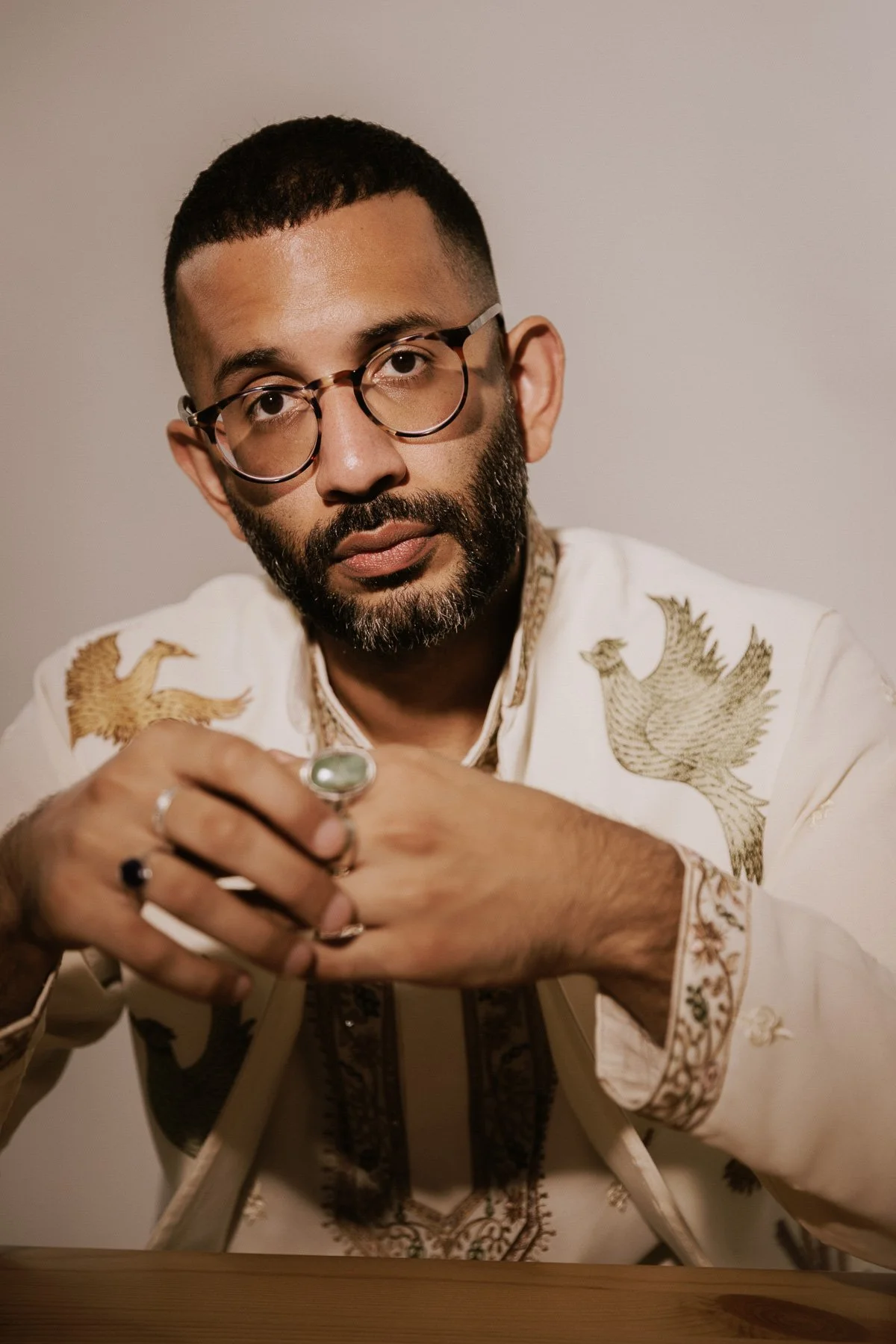 A man with glasses and a beard, wearing a white embroidered shirt, sitting at a table with hands clasped.