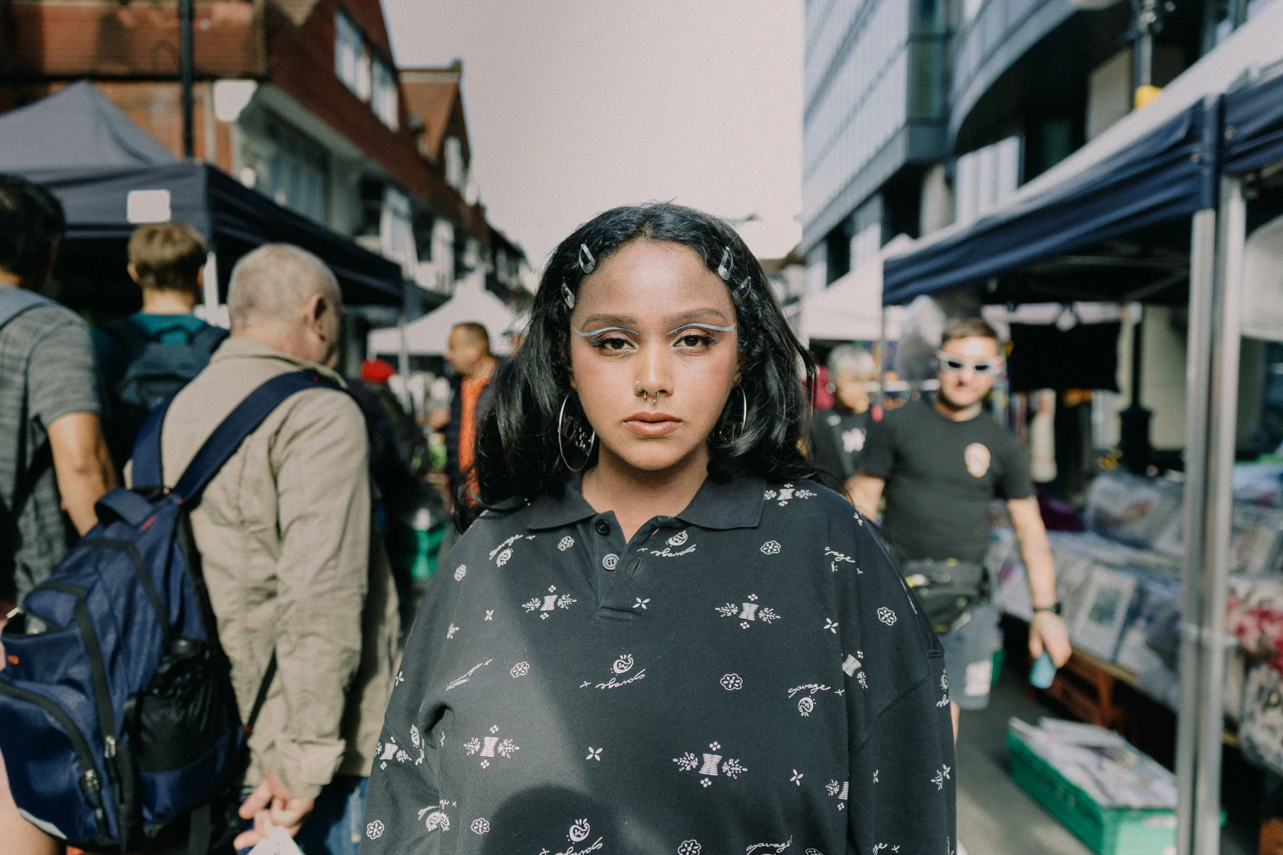 A young woman with black hair and light blue eye makeup standing in a bustling outdoor market.