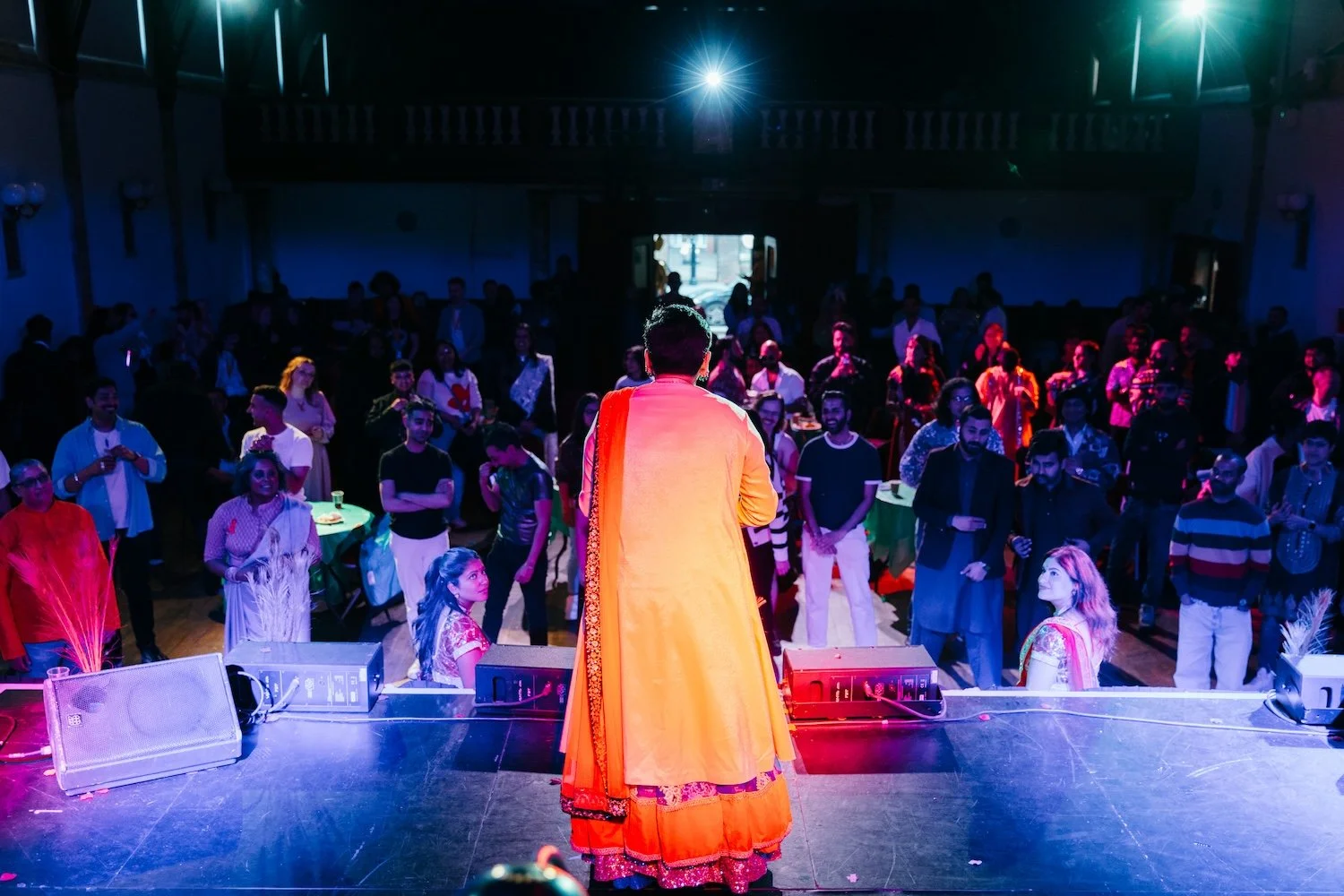Performer in traditional orange attire on stage facing audience, in a large hall with a crowd of people watching, colorful lighting, and a bright spotlight above.