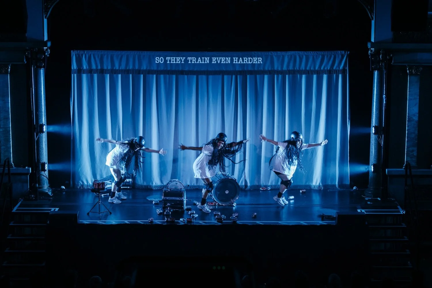 Three girls wearing helmets and white dresses performing a dance on stage with blue lighting, drums, and a blue curtain background that has the text 'SO THEY TRAIN EVEN HARDER' at the top.