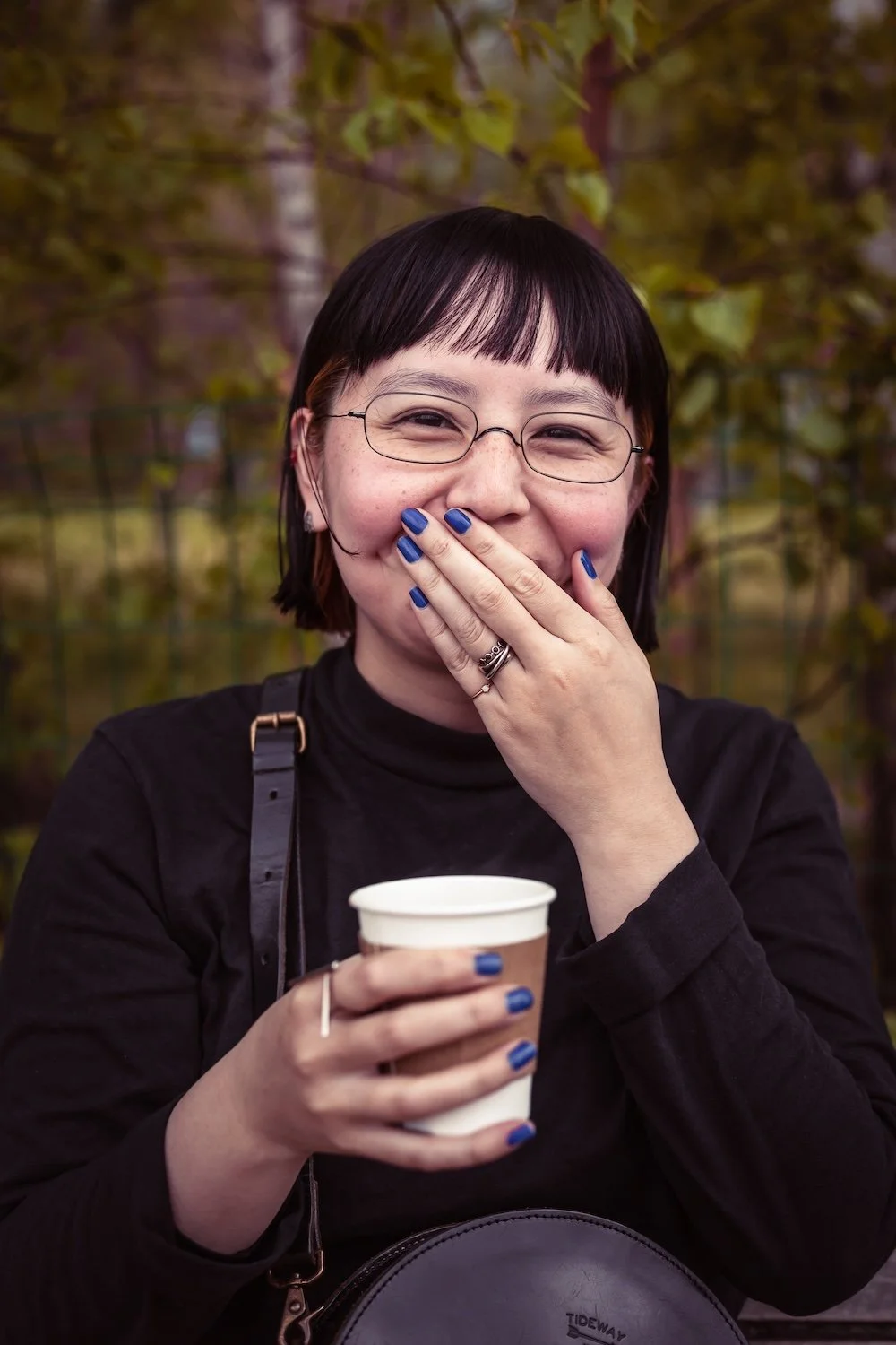 A woman with short dark hair and glasses smiling and covering her mouth with her hand, holding a disposable coffee cup in her other hand, outdoors with trees in the background.