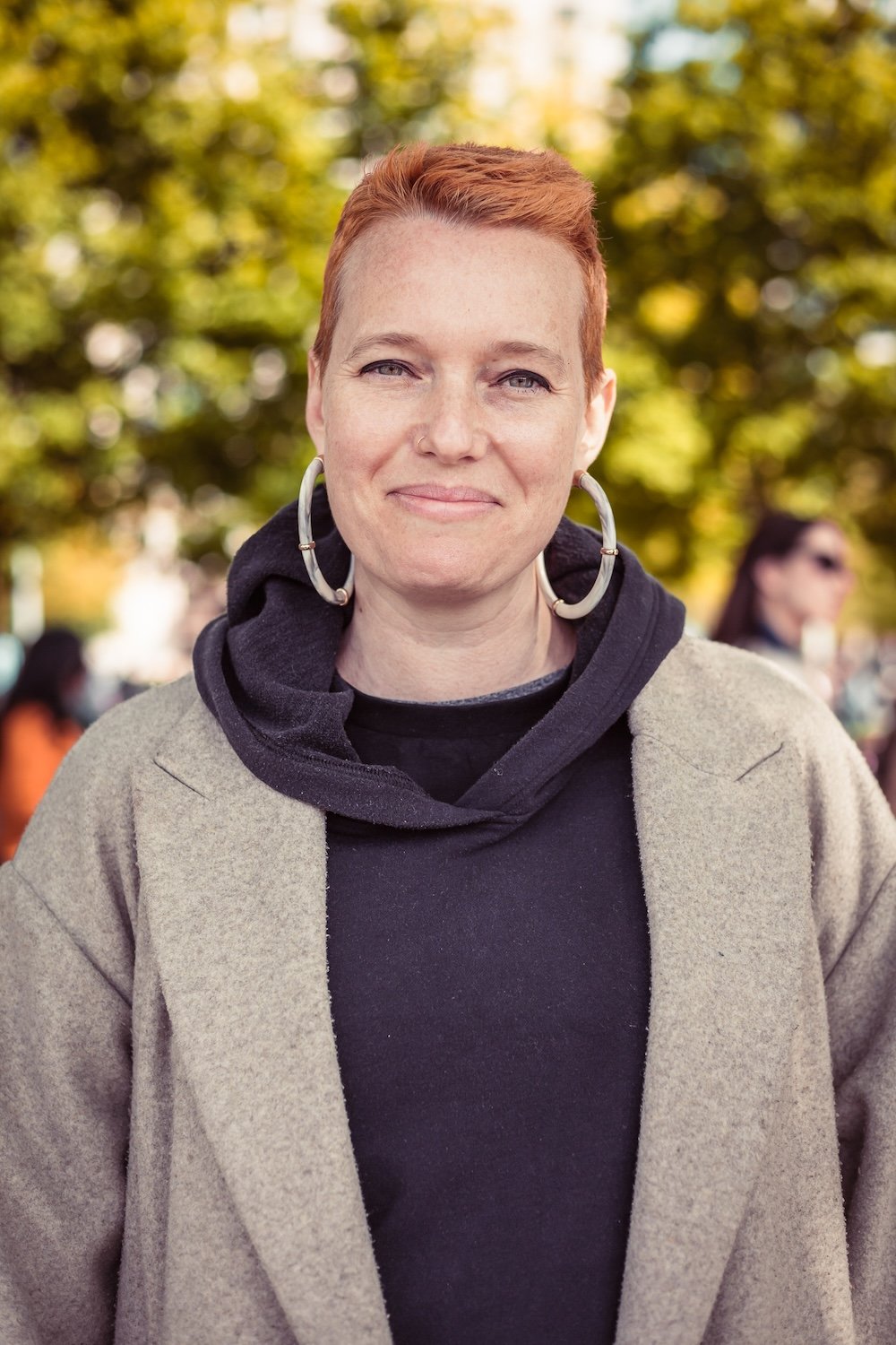 A woman with short red hair and hoop earrings standing outdoors with a background of trees and people, wearing a black hoodie and a beige coat.