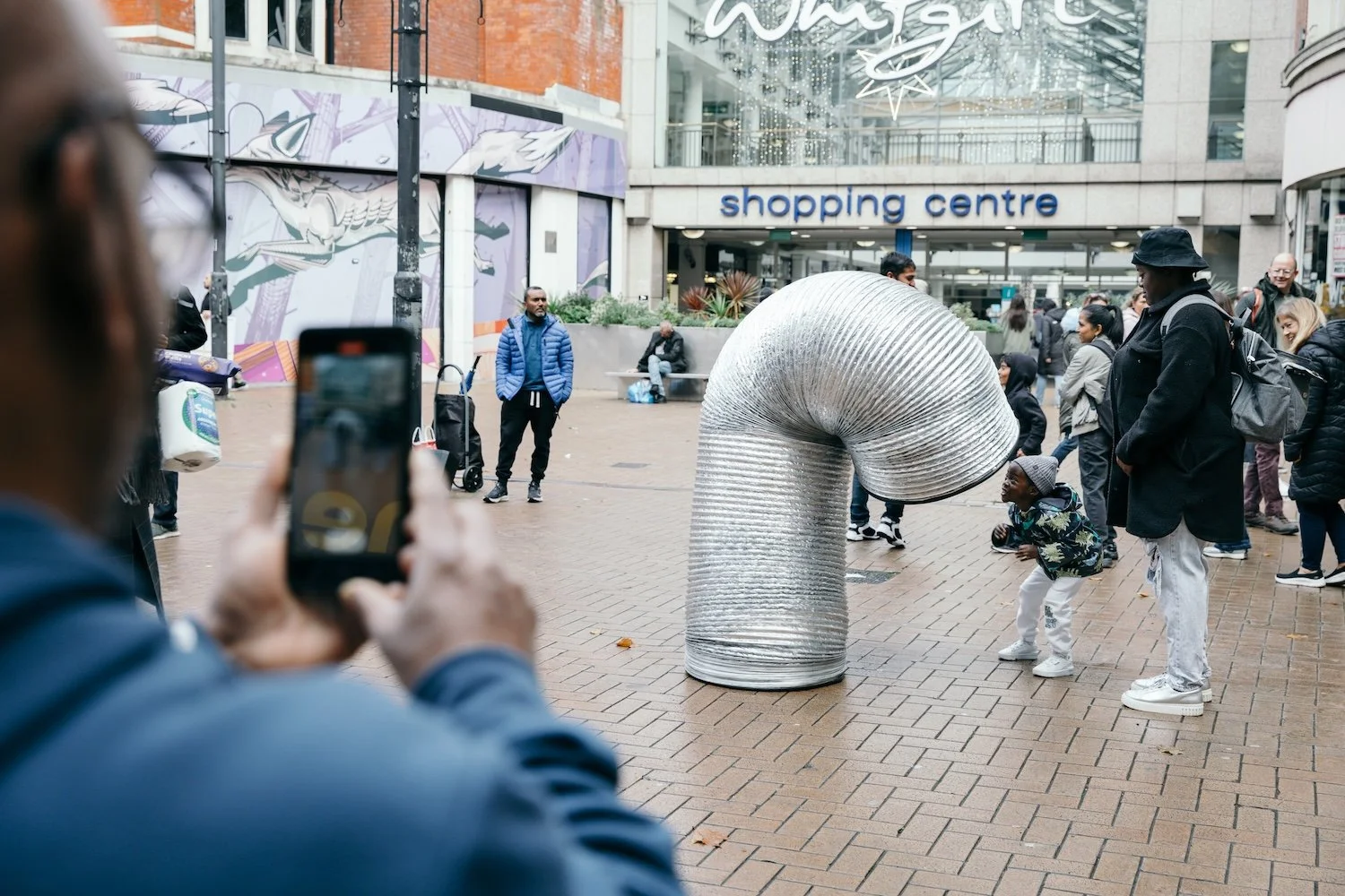 A street performer in a shiny, flexible, metallic tube costume bends over to interact with a young boy in a busy shopping area. People watch and take photos near a shopping center entrance, with some individuals standing and others sitting on benches