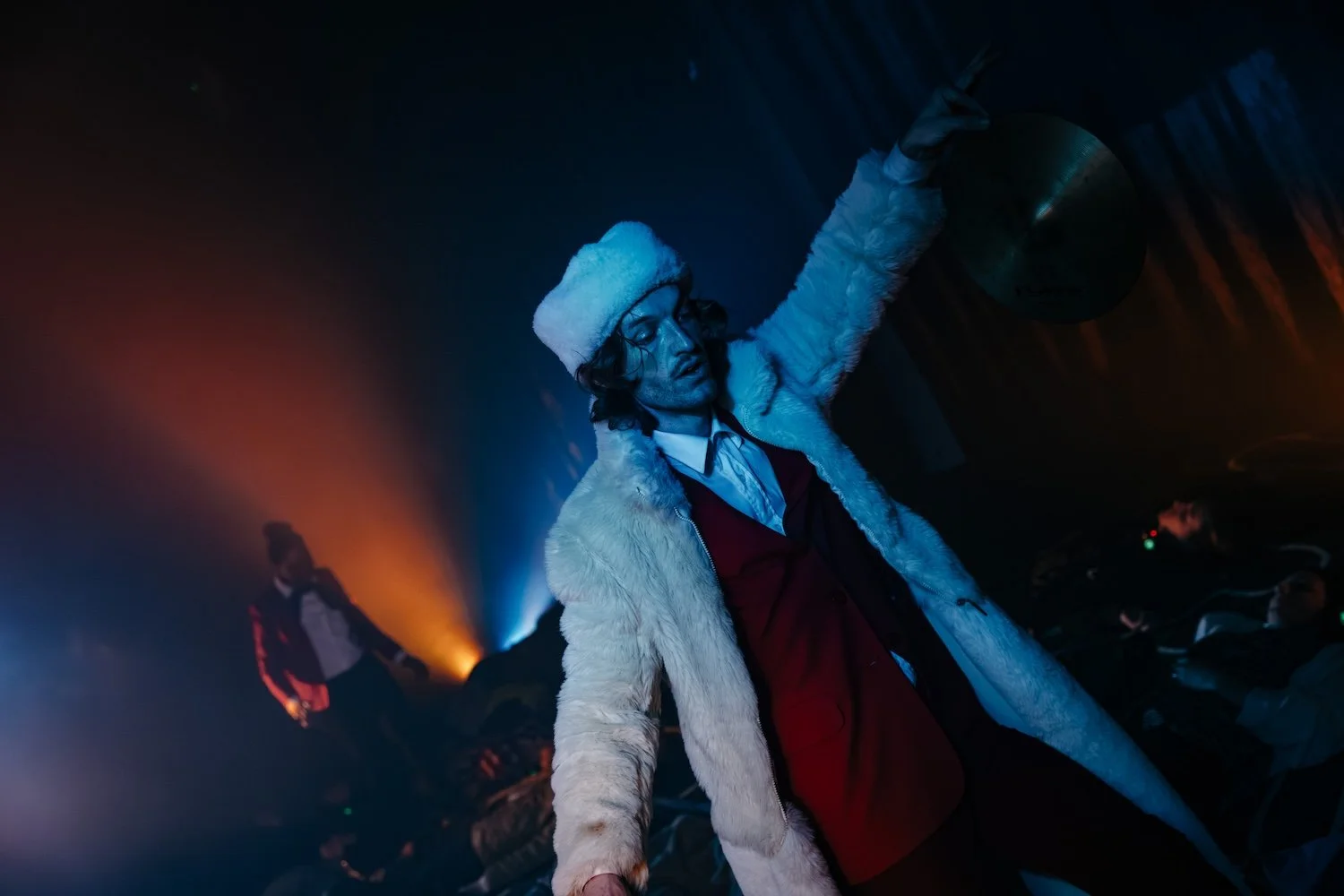 Person wearing a fur coat and hat, with long hair, at a dark party or club, with colorful lighting and other people in the background.