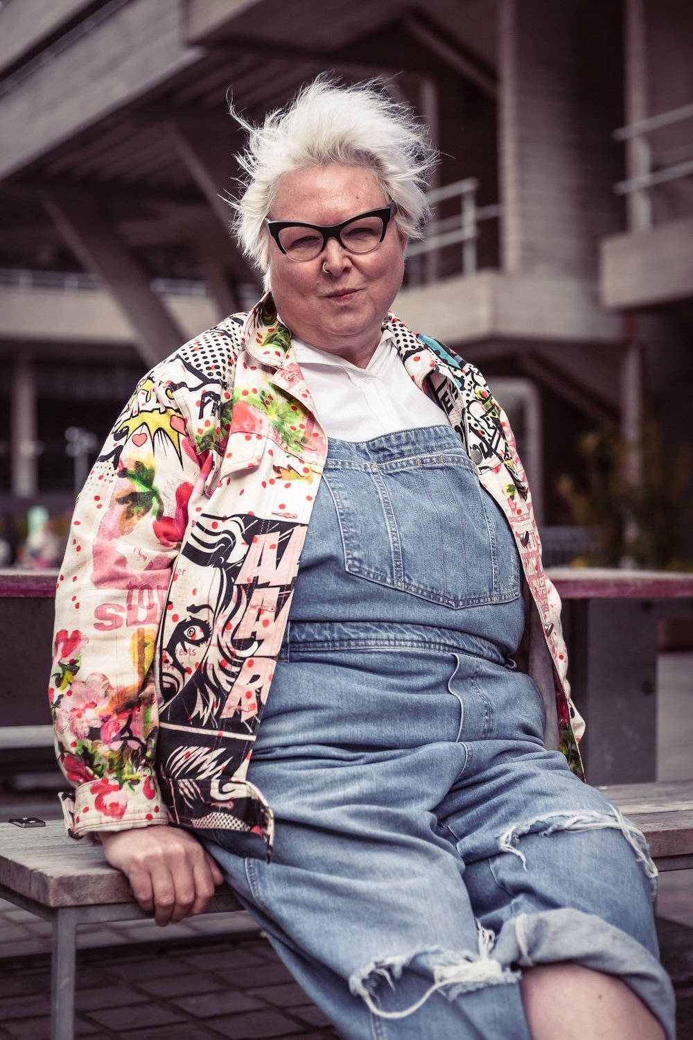 A woman with short, spiky white hair, wearing black-rimmed glasses, a colorful patterned jacket, a white shirt, and ripped jeans, sitting on a bench outdoors.