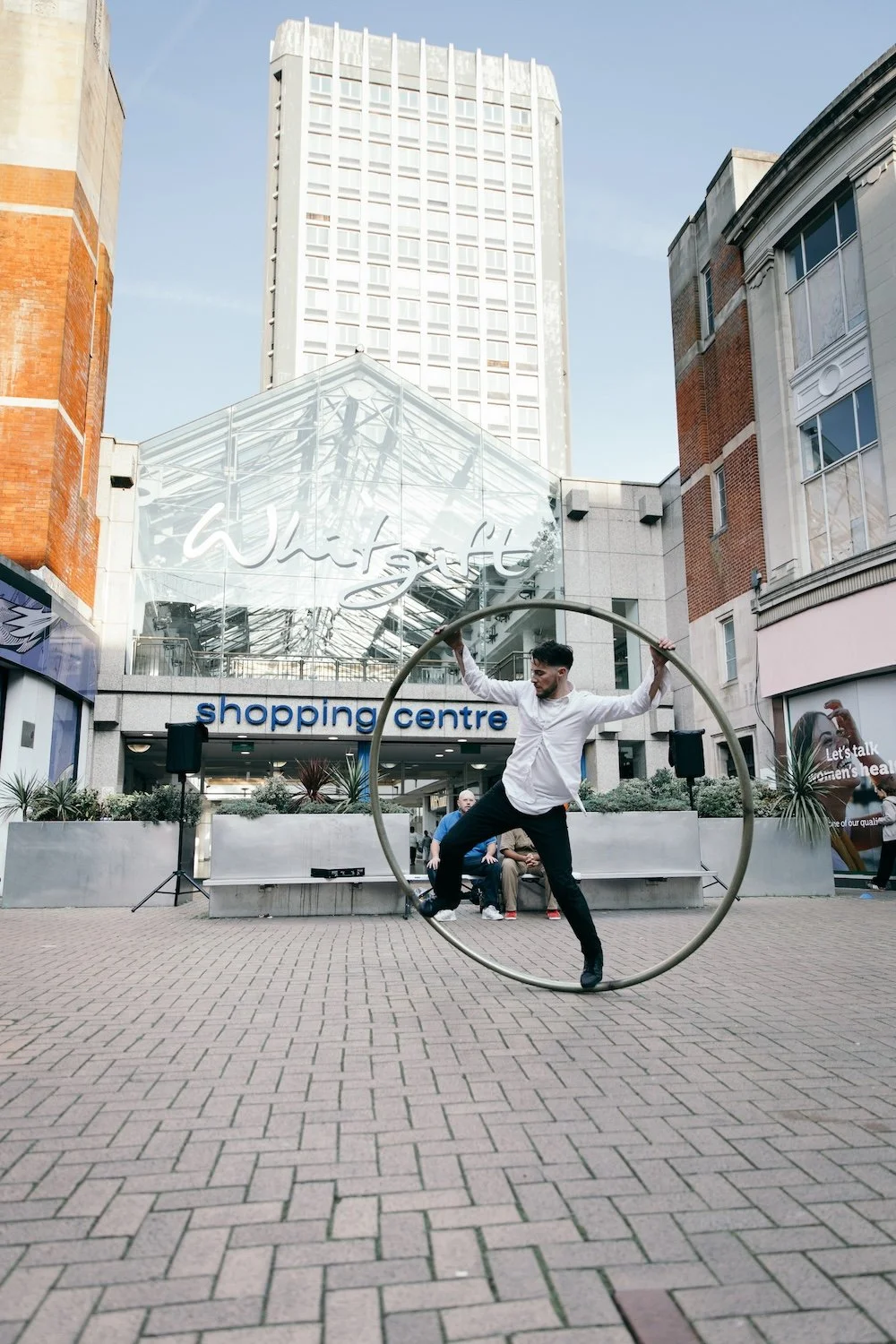 A man performing a trick on a wheel in front of a shopping centre with a glass arch entrance, surrounded by buildings and a few spectators seated nearby.