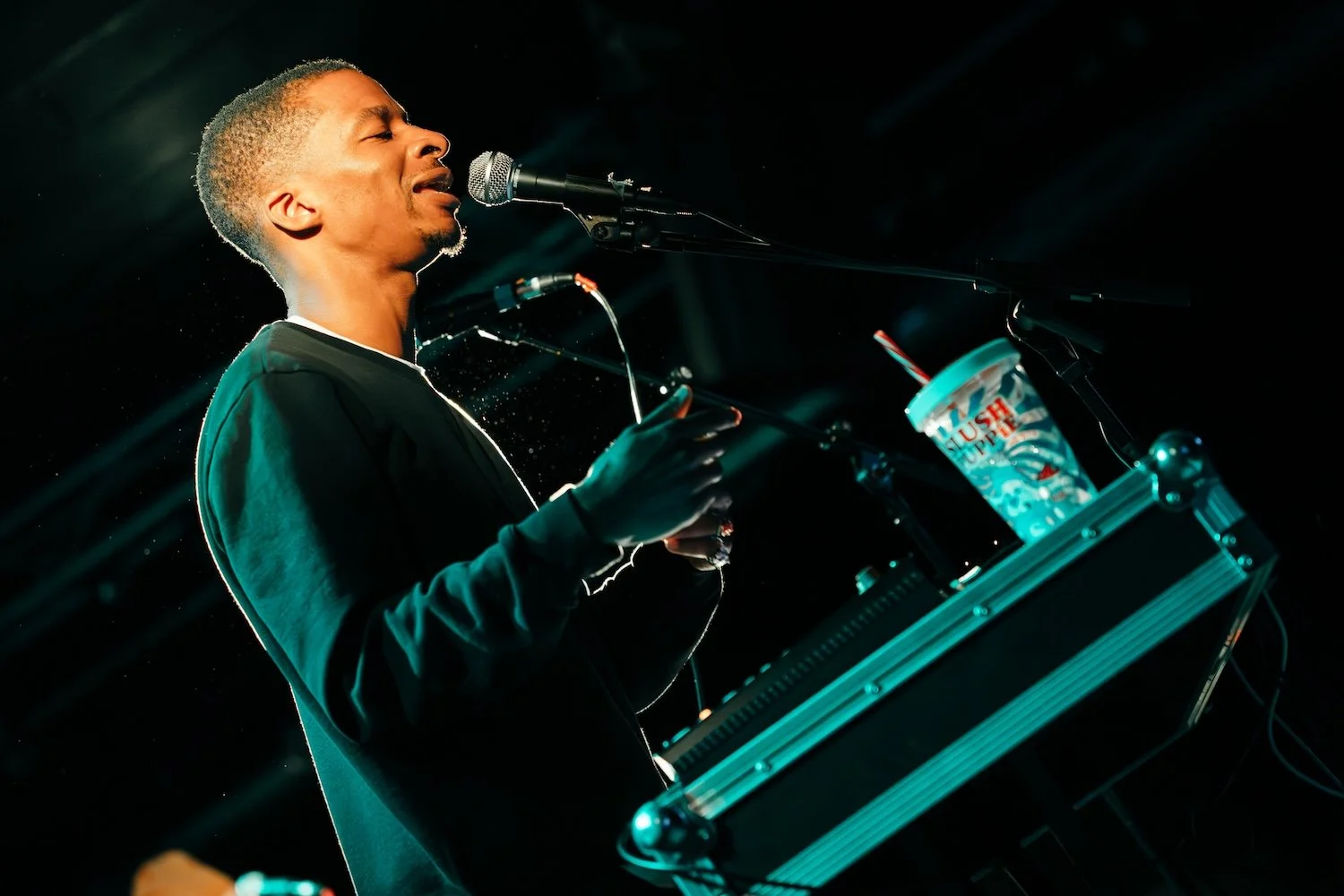 A male musician performing on stage singing into a microphone with a drink cup nearby.