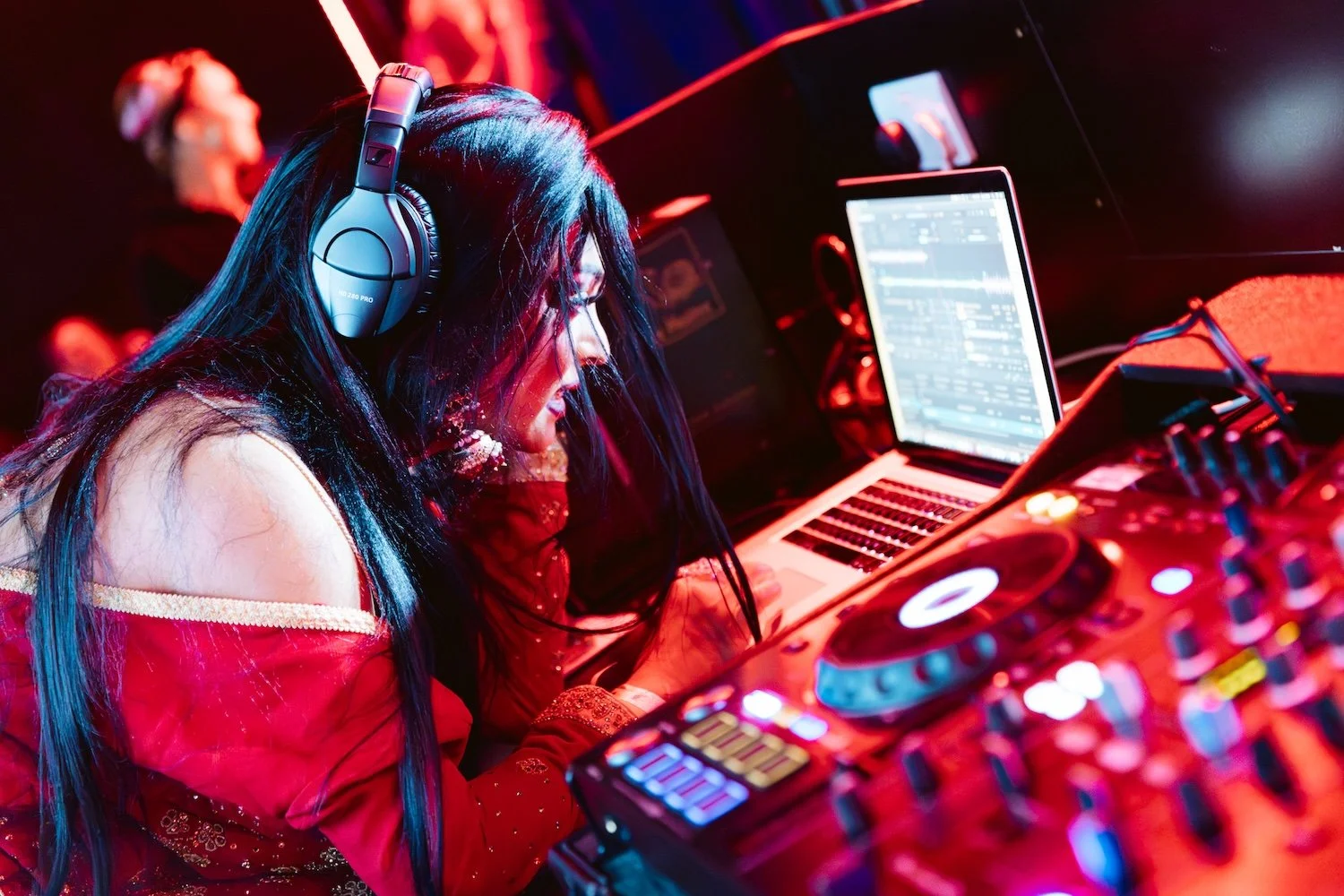 A female DJ with long dark hair wearing headphones, operating DJ equipment and a laptop in a dark, colorful nightclub setting.