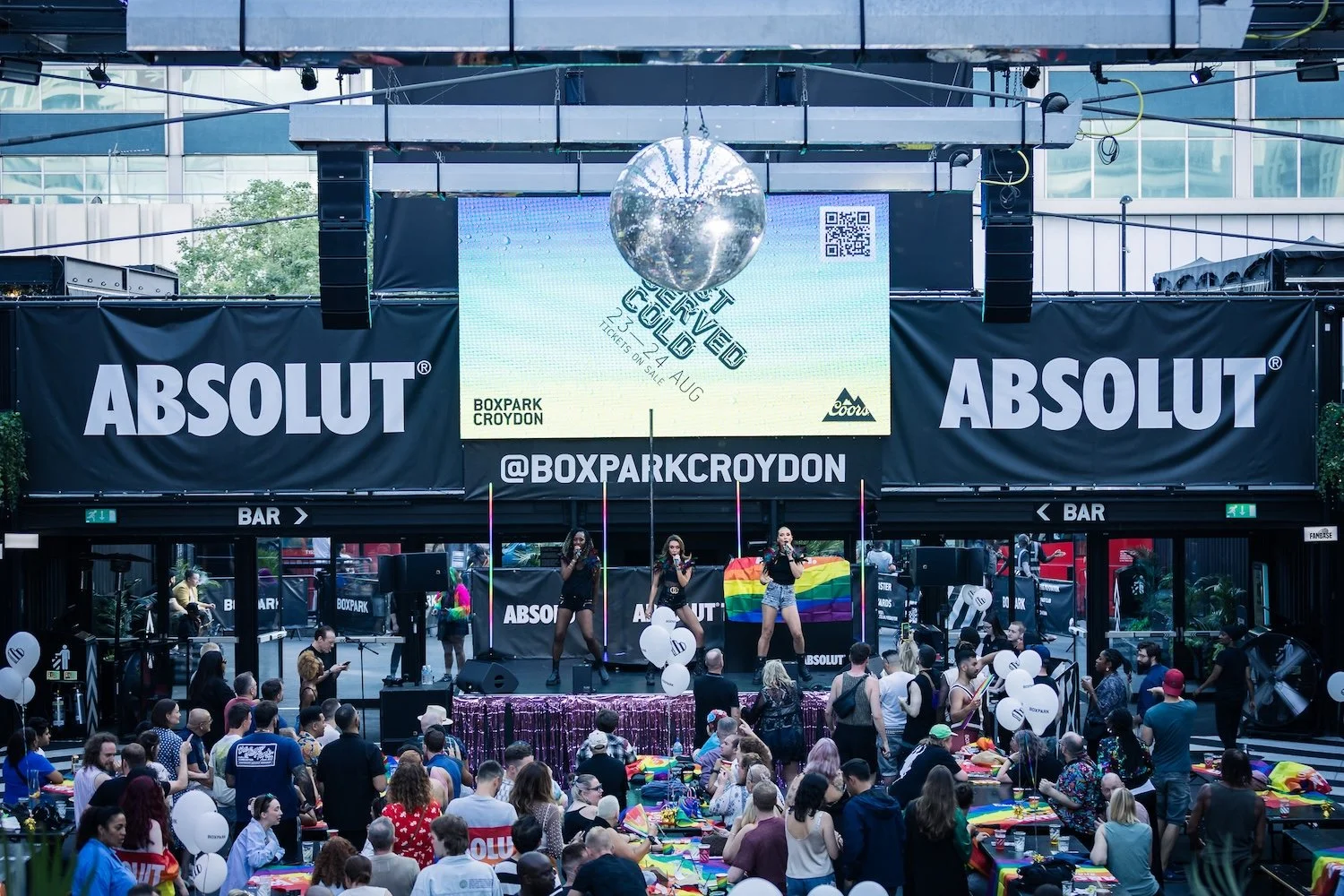 Crowd attending a celebration event at Boxpark Croydon with a large stage, performers, and rainbow flags, promoting inclusivity and pride, sponsored by Absolut.
