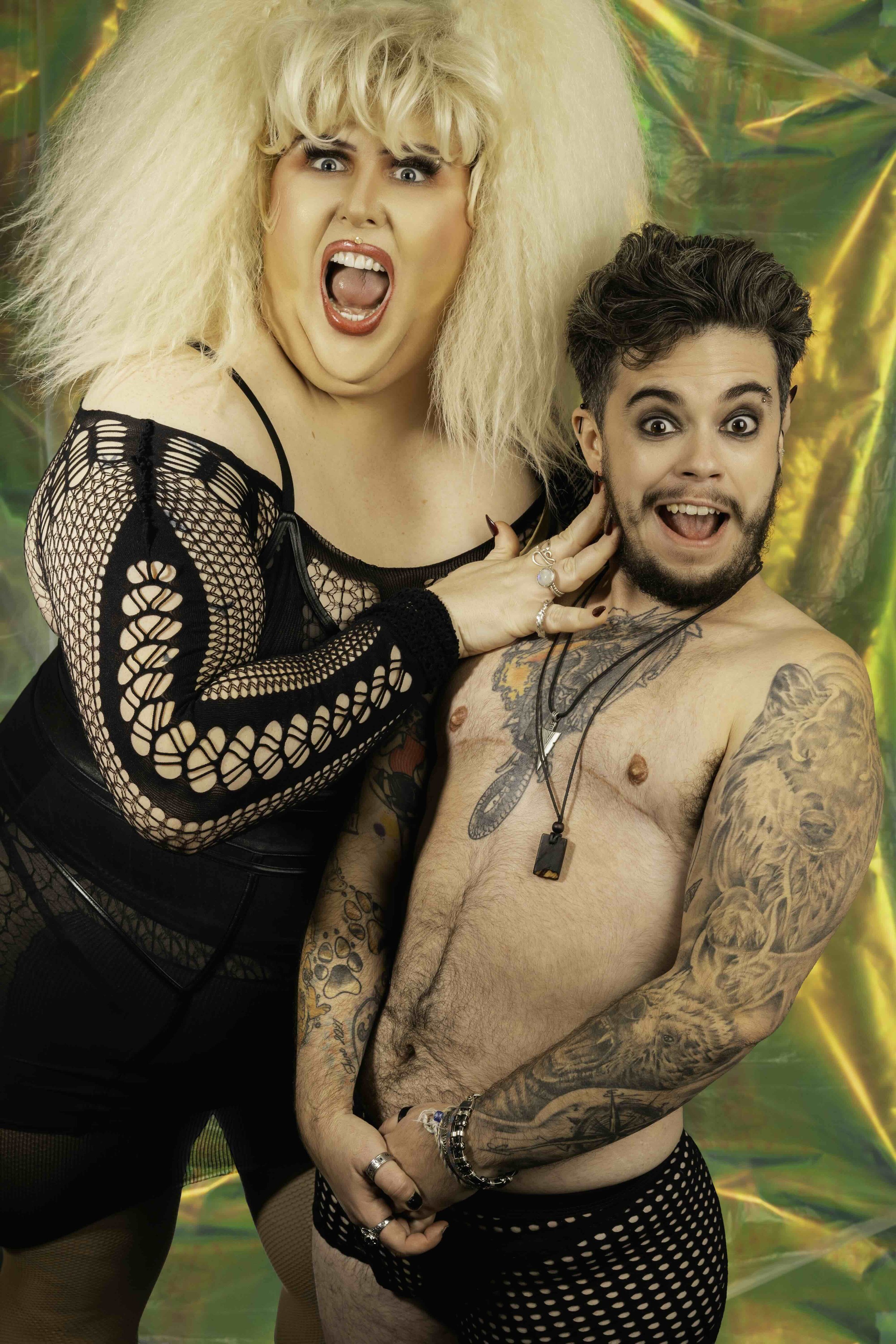 A woman with blond, frizzy hair and makeup, wearing a black fishnet top, touching a shirtless man with tattoos, wearing polka dot underwear, against a colorful, iridescent background.