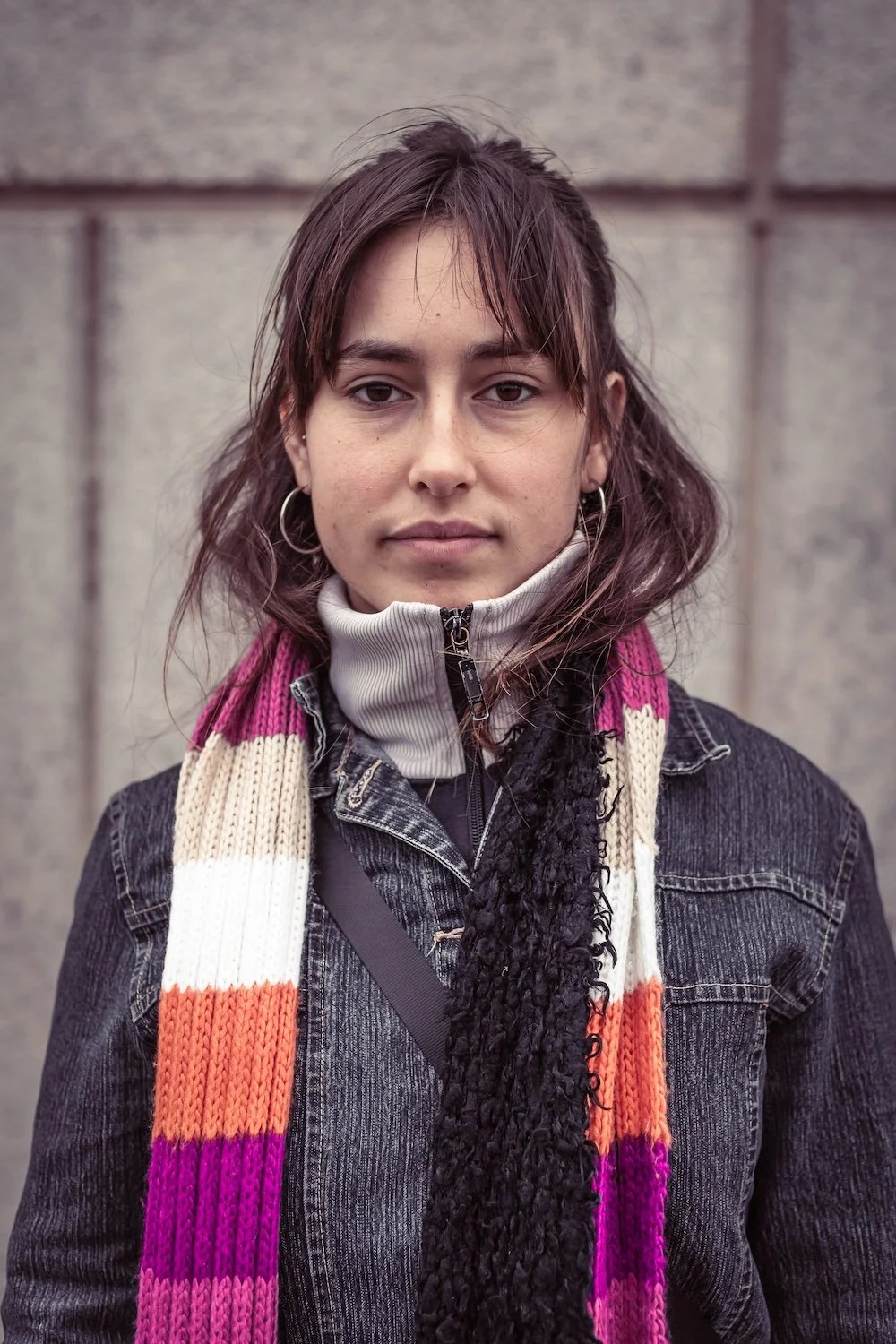 A young woman with shoulder-length brown hair, wearing a black denim jacket, a rainbow-colored scarf, and a light gray zip-up turtleneck, standing outdoors against a gray stone wall.