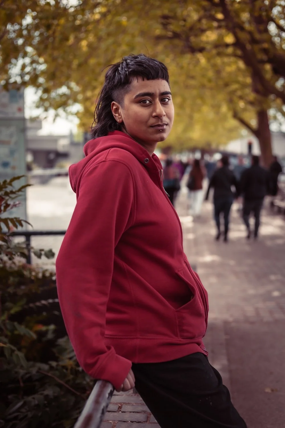A person with short dark hair wearing a red hoodie standing outdoors on a sidewalk, with trees and people walking in the background during autumn.