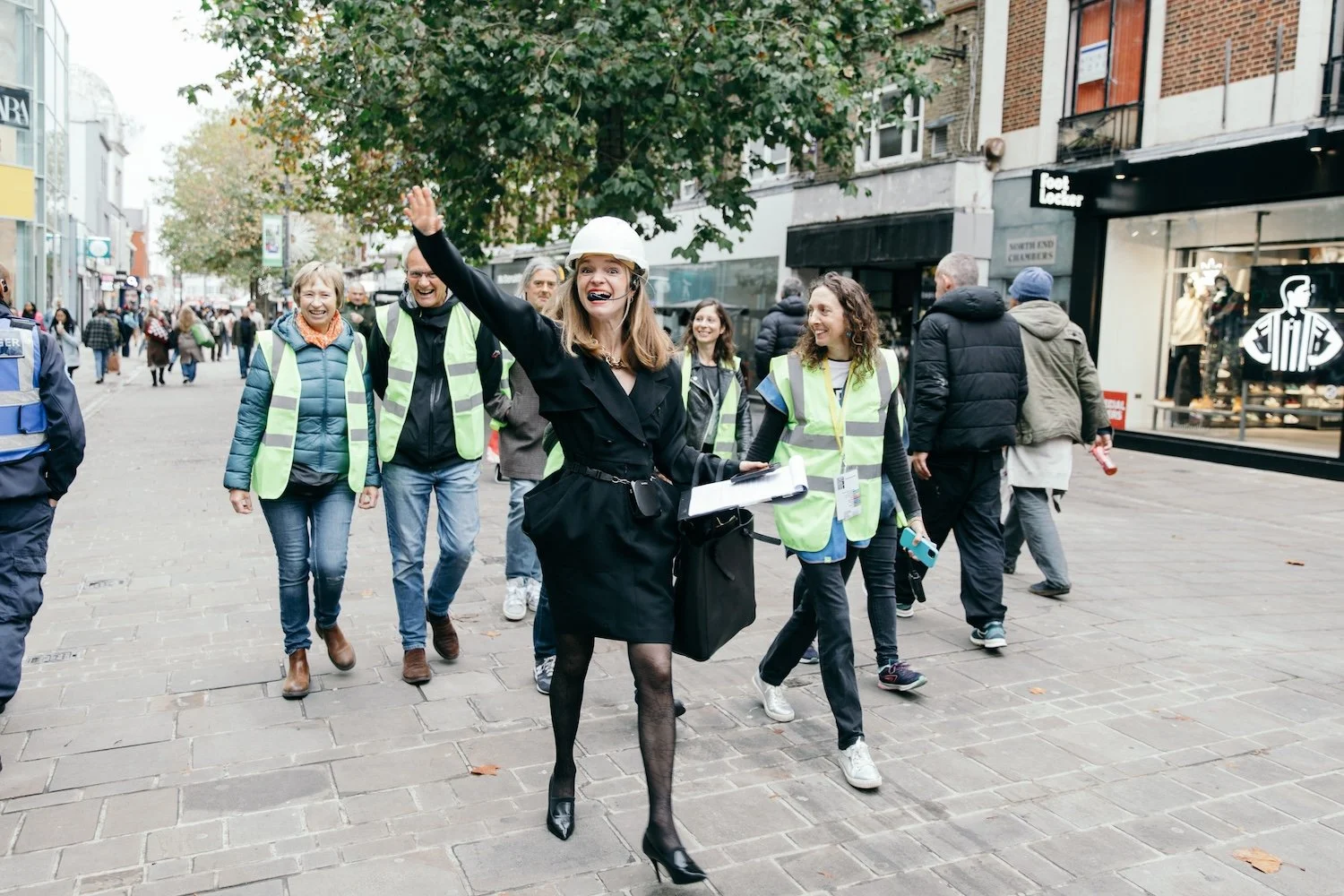 Group of people walking on a city sidewalk, with a woman in a black dress and construction helmet leading and smiling, surrounded by others wearing high-visibility vests, some holding face masks, during daytime.