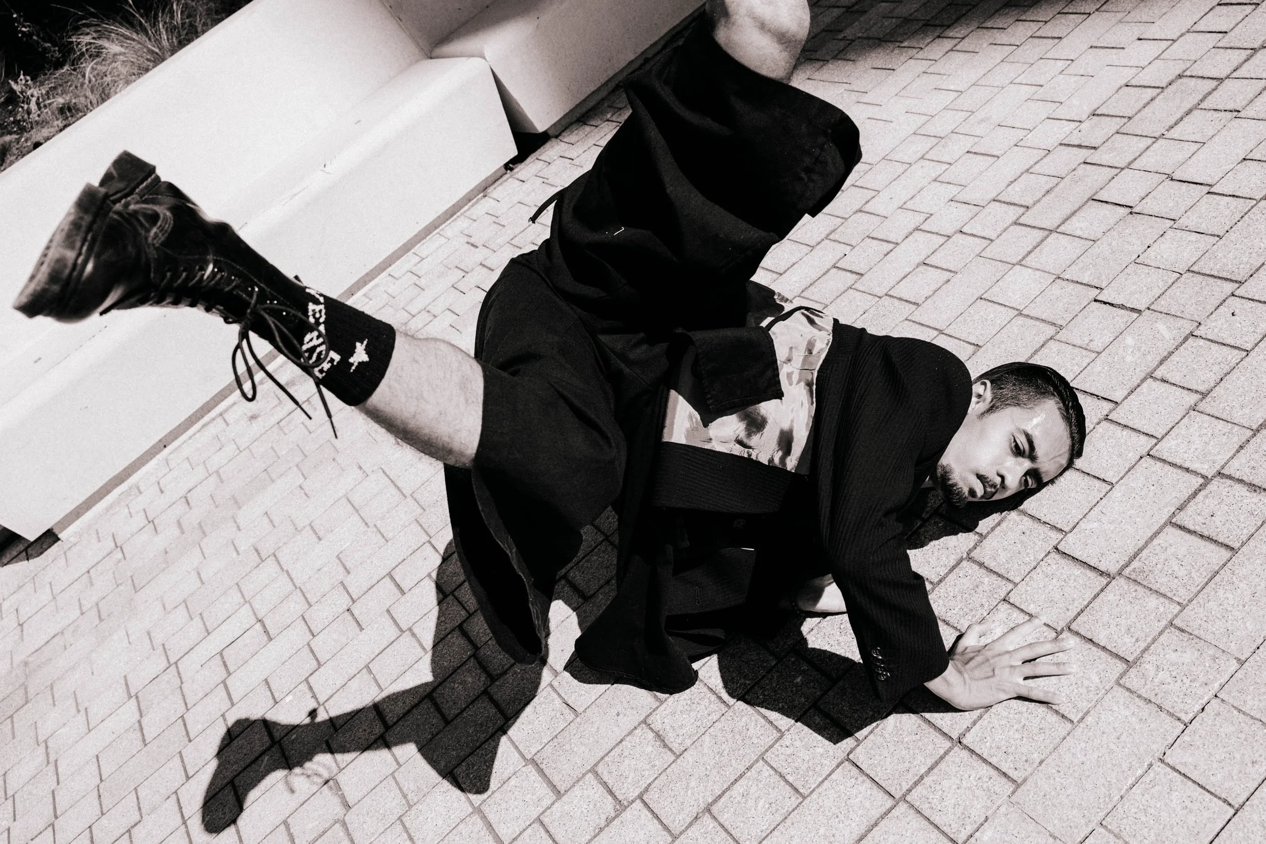 A man dressed in dark clothing, including a blazer, camouflage T-shirt, and black sports socks, is lying on a brick sidewalk. He is on his hands and knees with his face turned to one side, and appears to be looking at the camera. 