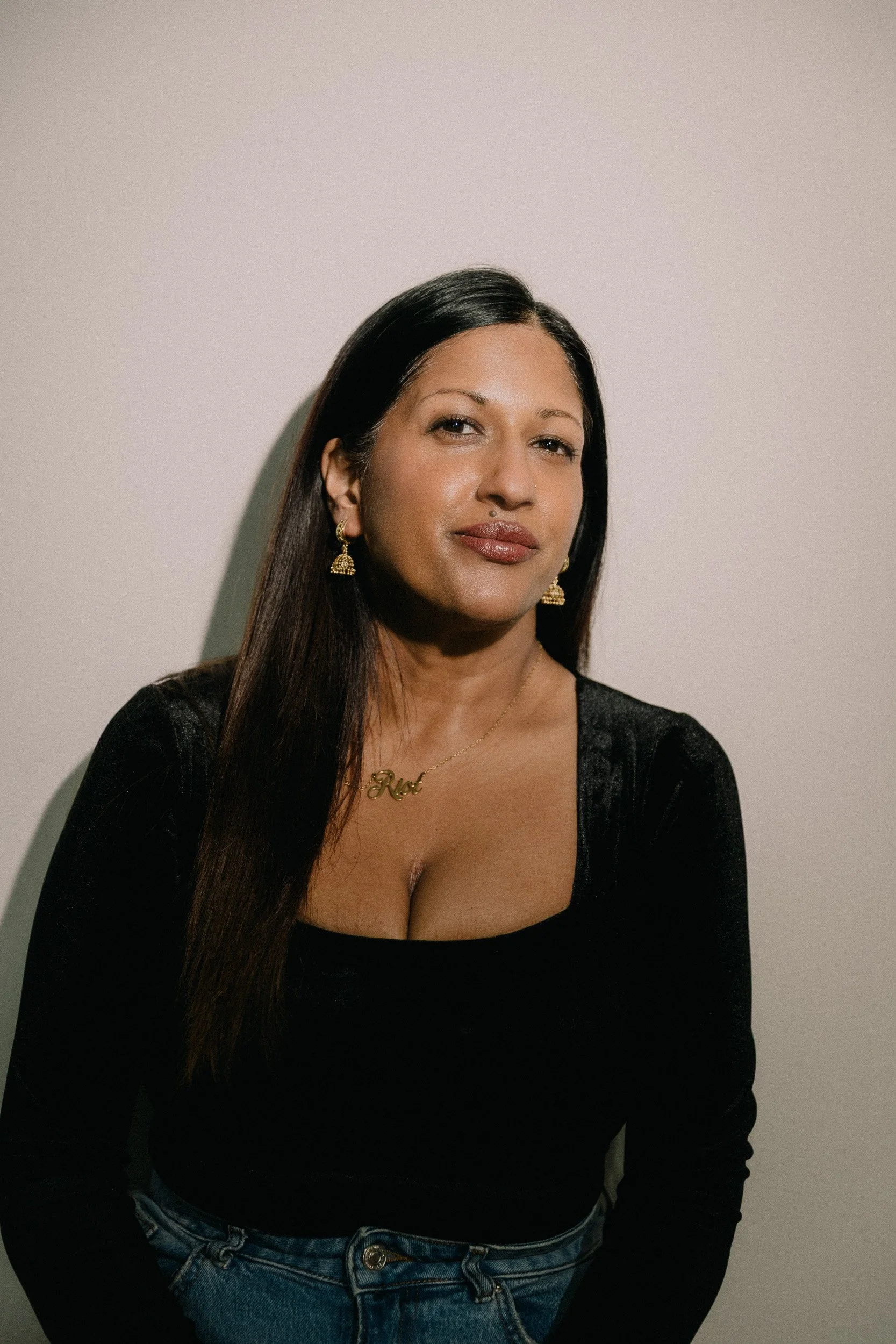 A woman with long black hair wearing gold earrings, a gold necklace, and a black top with jeans standing against a plain wall.