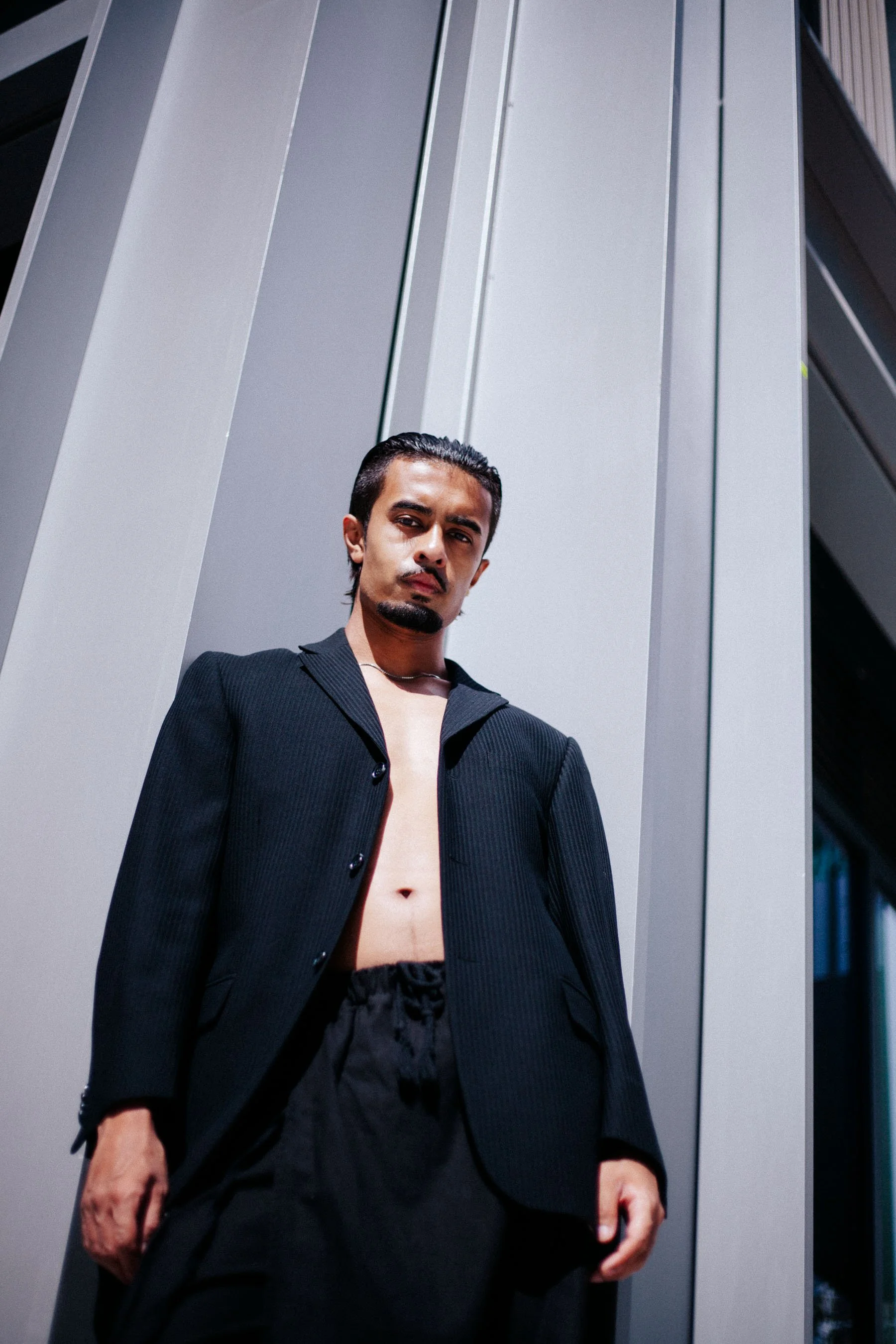 A young man with slicked-back hair, a mustache and goatee, wearing a black blazer and black shorts, posing against a modern, metallic building exterior under bright sunlight.