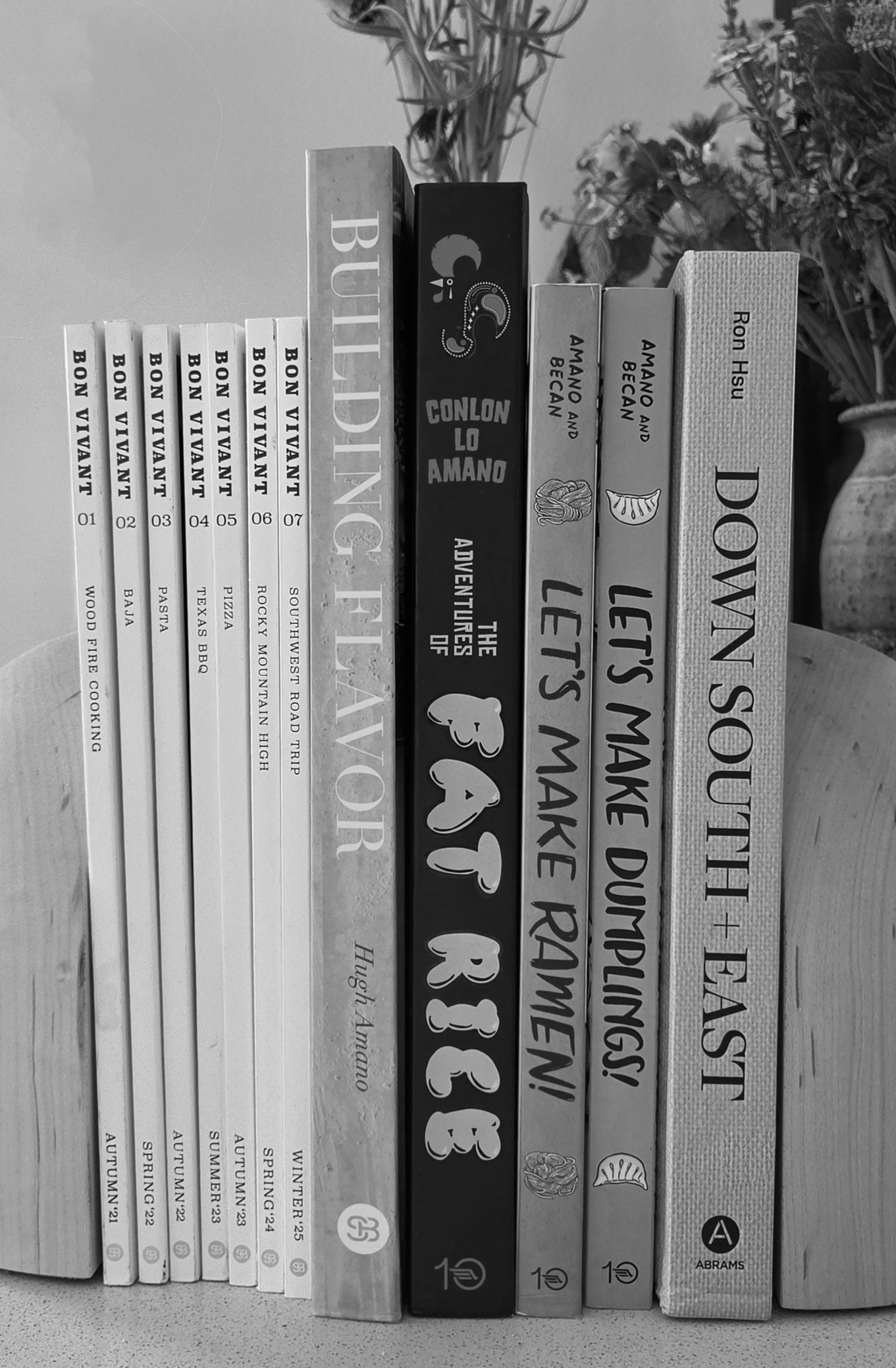 Stacked books on a surface with titles related to cooking, travel, and food, including 'Bon Vivant,' 'The Adventures of Fat Rice,' and 'Let's Make Ramen!,' with some decorative items and plants in the background.
