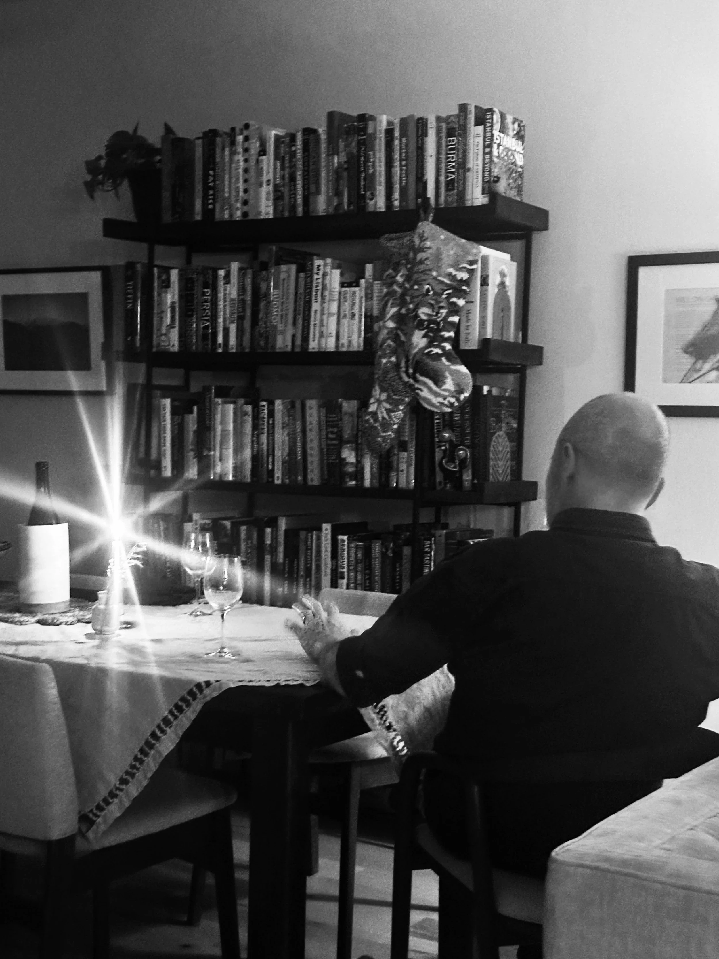 A man with a bald head sitting at a dining table with a tablecloth, wine glasses, and a candle, facing a bookshelf filled with books and a framed picture on the wall.