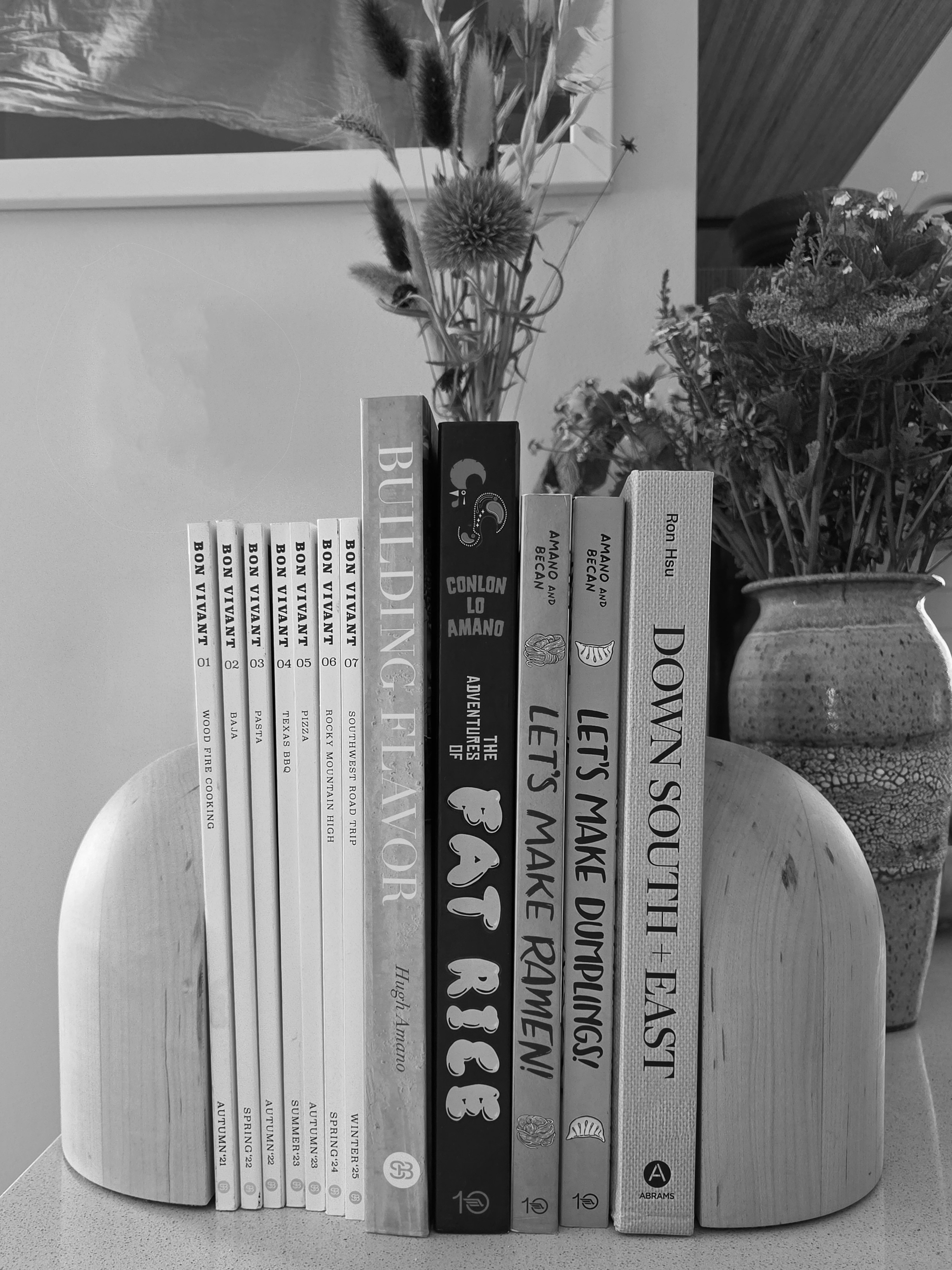 A shelf with a row of seven white books titled 'BON VIVANT,' each numbered 01 to 07, and three books with titles ''Down South + East", 'LET'S MAKE DUMPLINGS,' and 'THE ADVENTURES OF Fat Rice'