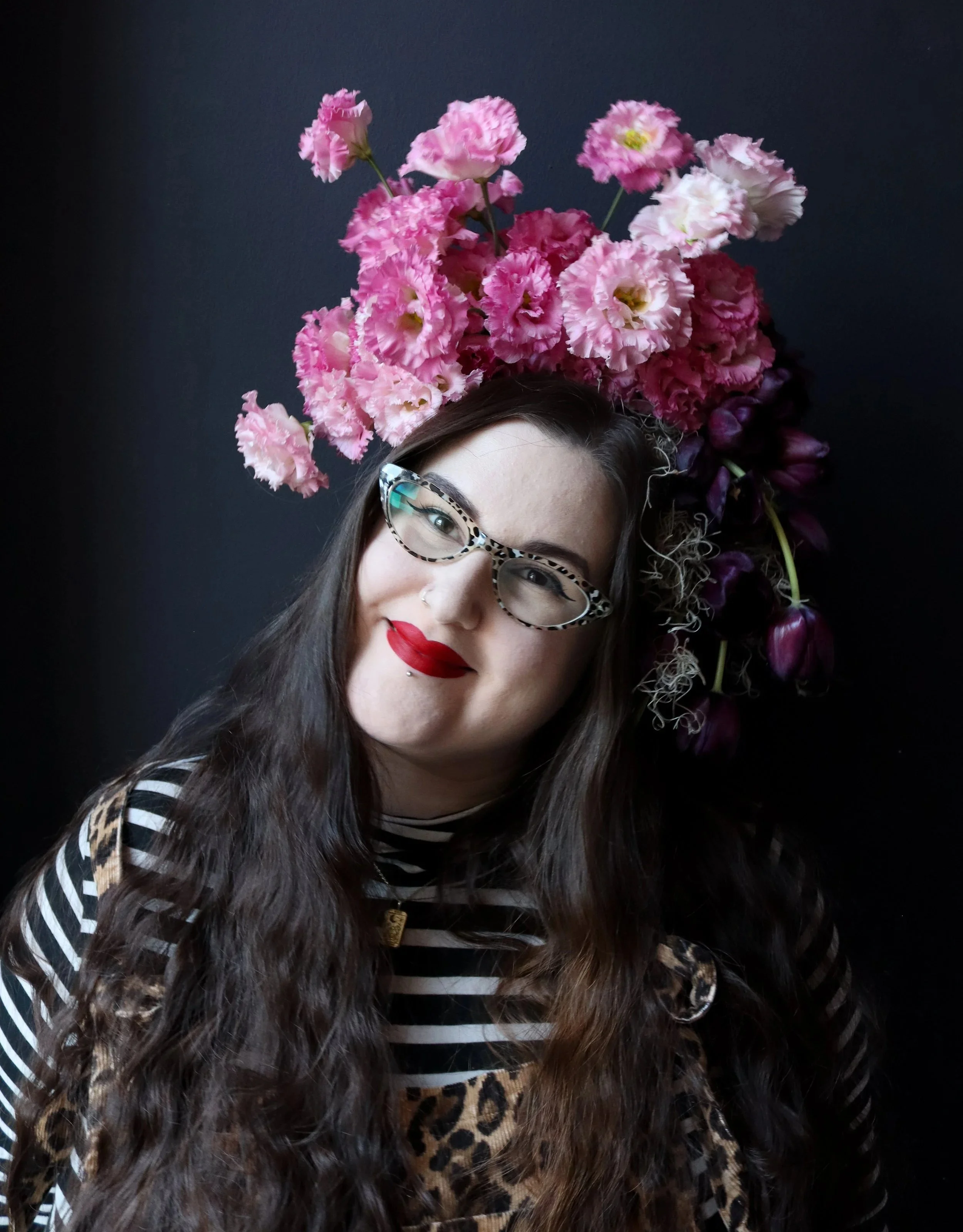A woman with long wavy hair, red lipstick, wearing leopard print glasses, a nose piercing, a striped shirt, and leopard print overalls, standing against a dark background with a floral arrangement on her head.