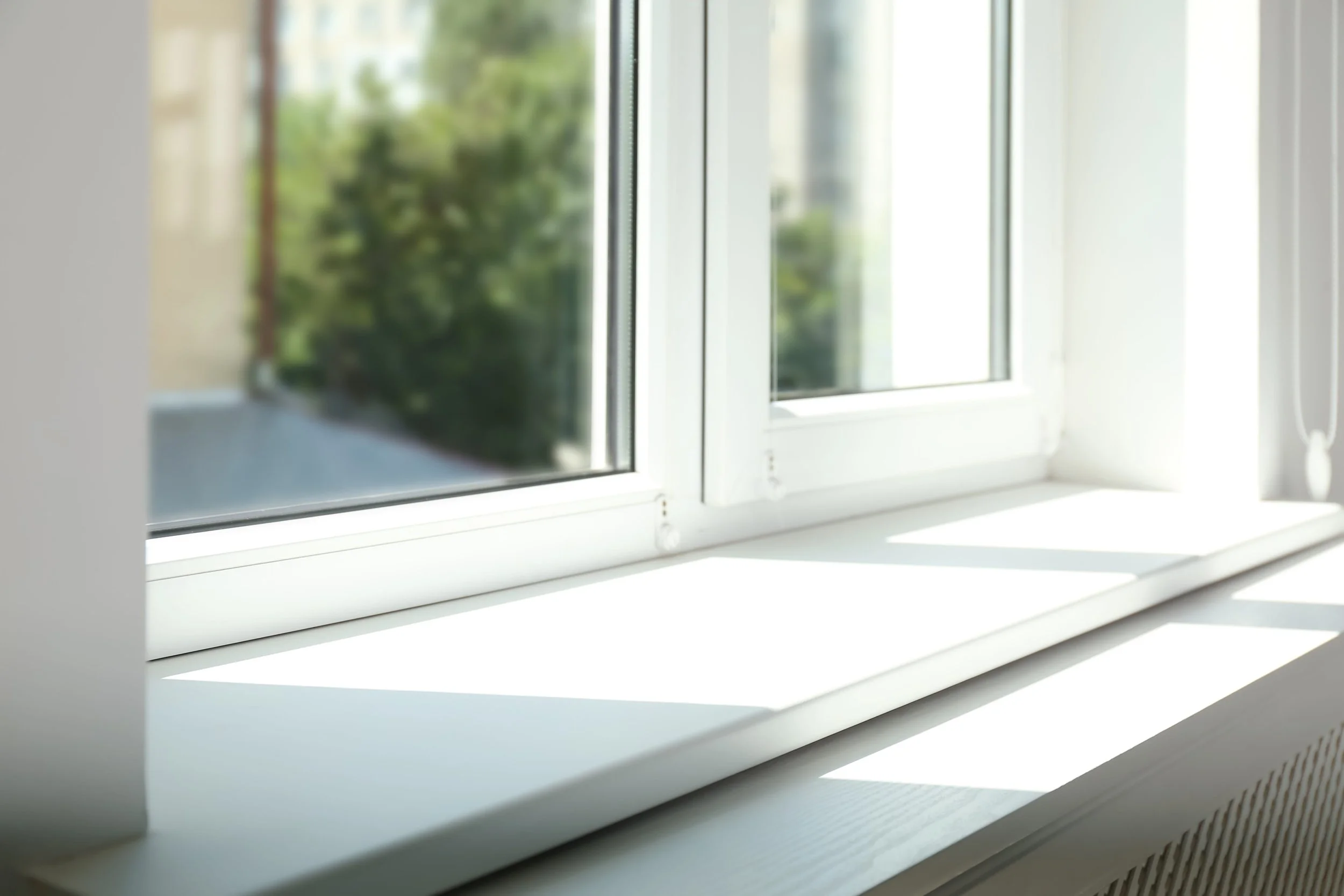 Sunlit white windowsill with shadows from a bright window frame. Blurred background of greenery and buildings outside. Airy and serene ambiance.